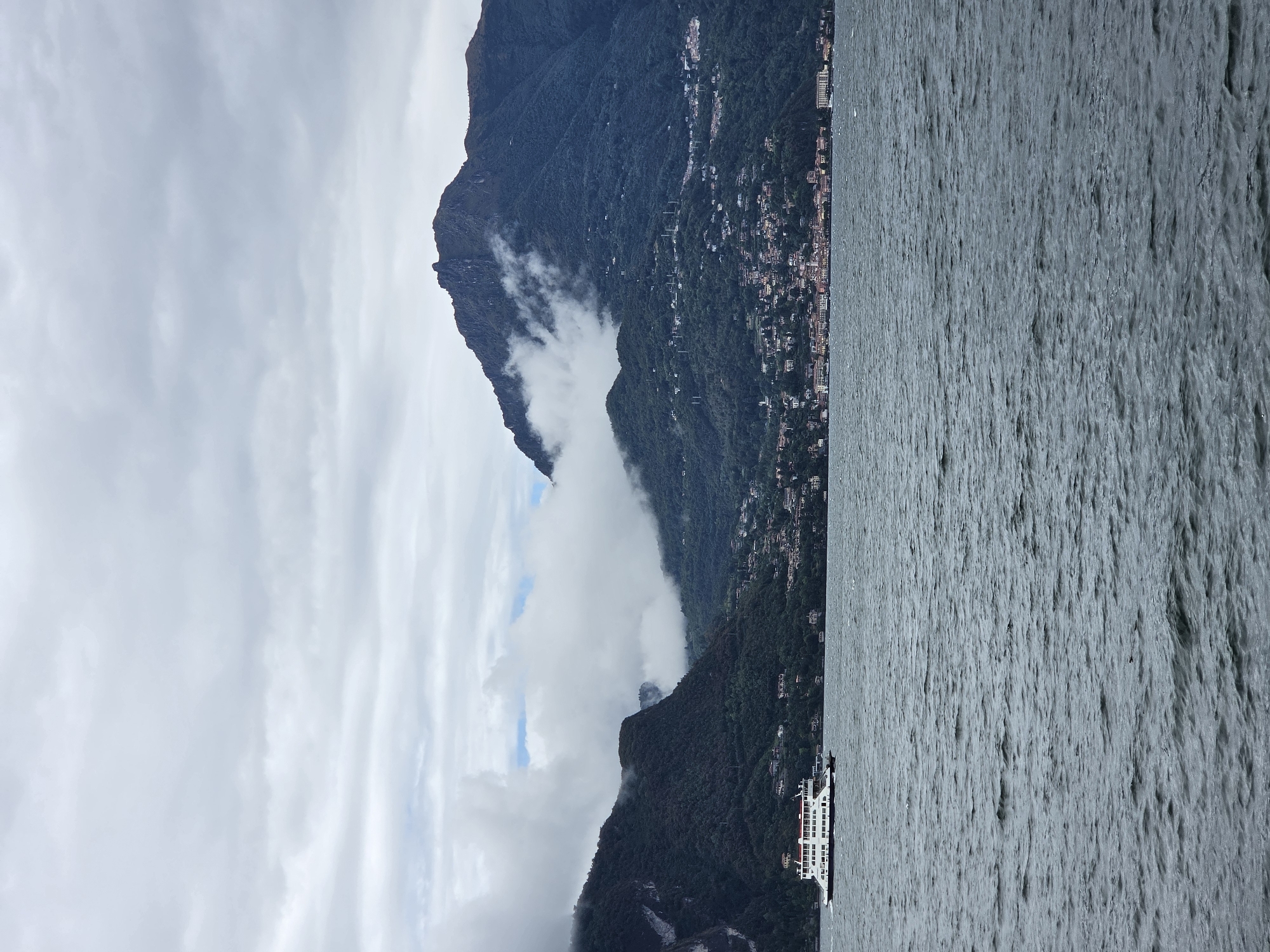 The image depicts a panoramic view of a body of water, likely a lake, with a ferryboat in the foreground. The ferry is white with a red roof and appears to be moving from left to right across the frame. In the background, a lush green mountainside rises steeply from the water. A town or village is nestled on the slope of the mountain, with buildings of various colors visible amongst the trees. The sky is overcast with a dramatic display of clouds, some of which are partially obscuring the upper reaches of the mountains. The water is choppy, with ripples and waves visible. The overall scene suggests a scenic, possibly European, location.