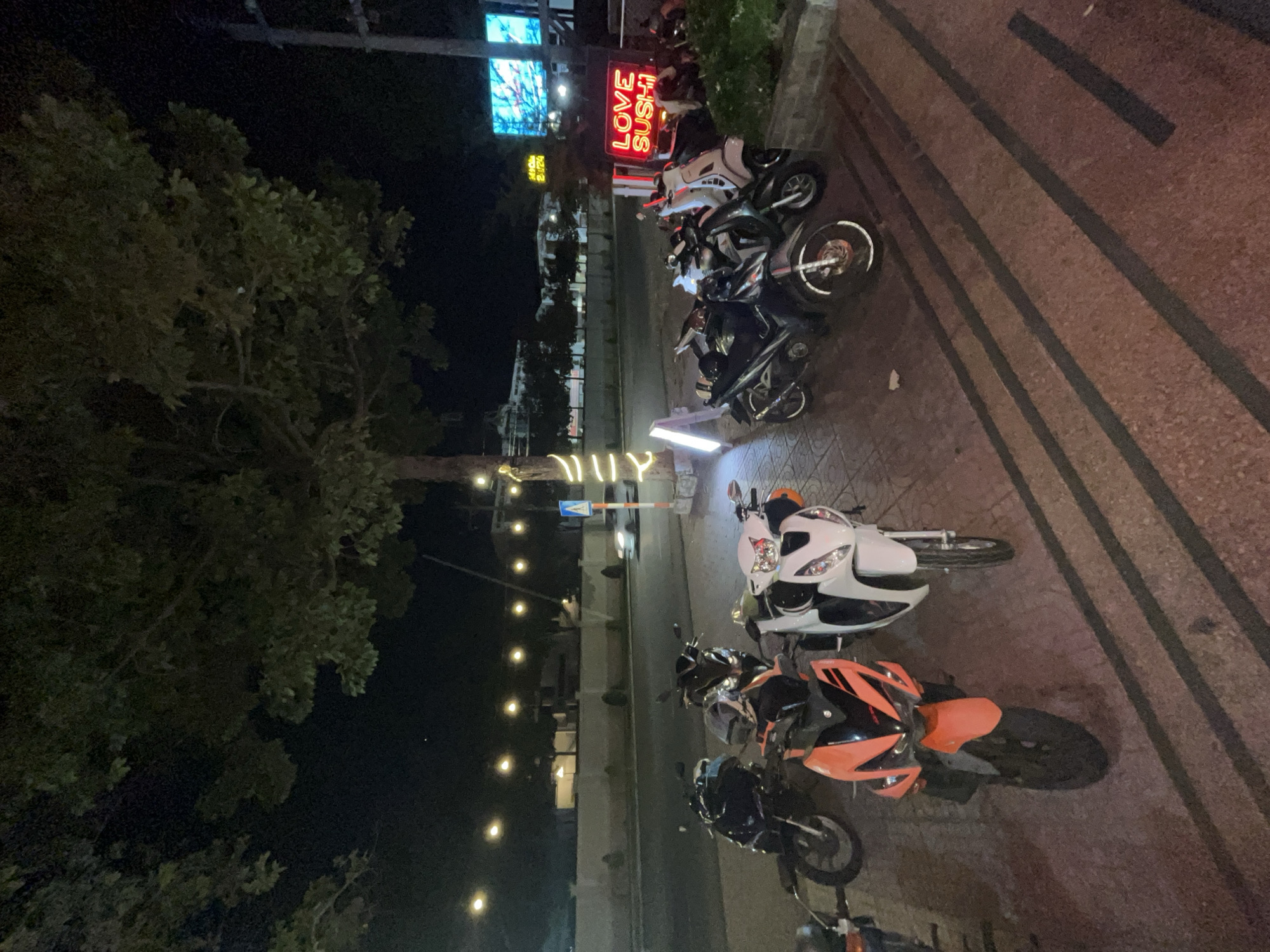 The image depicts a nighttime scene on a sidewalk in front of a business in Phu Thuy Ward, Vietnam. Several motorcycles are parked on the sidewalk. A neon sign that reads "LOVE SUSHI" is visible on the right side of the frame, illuminated in red. In the background, a street with traffic and buildings can be seen. The lighting suggests it is after dark, with streetlights and artificial lighting from the businesses casting illumination on the scene. A large tree with dark foliage is prominent in the upper portion of the image, with decorative lights strung along its trunk and branches. The ground is paved with textured tiles.
