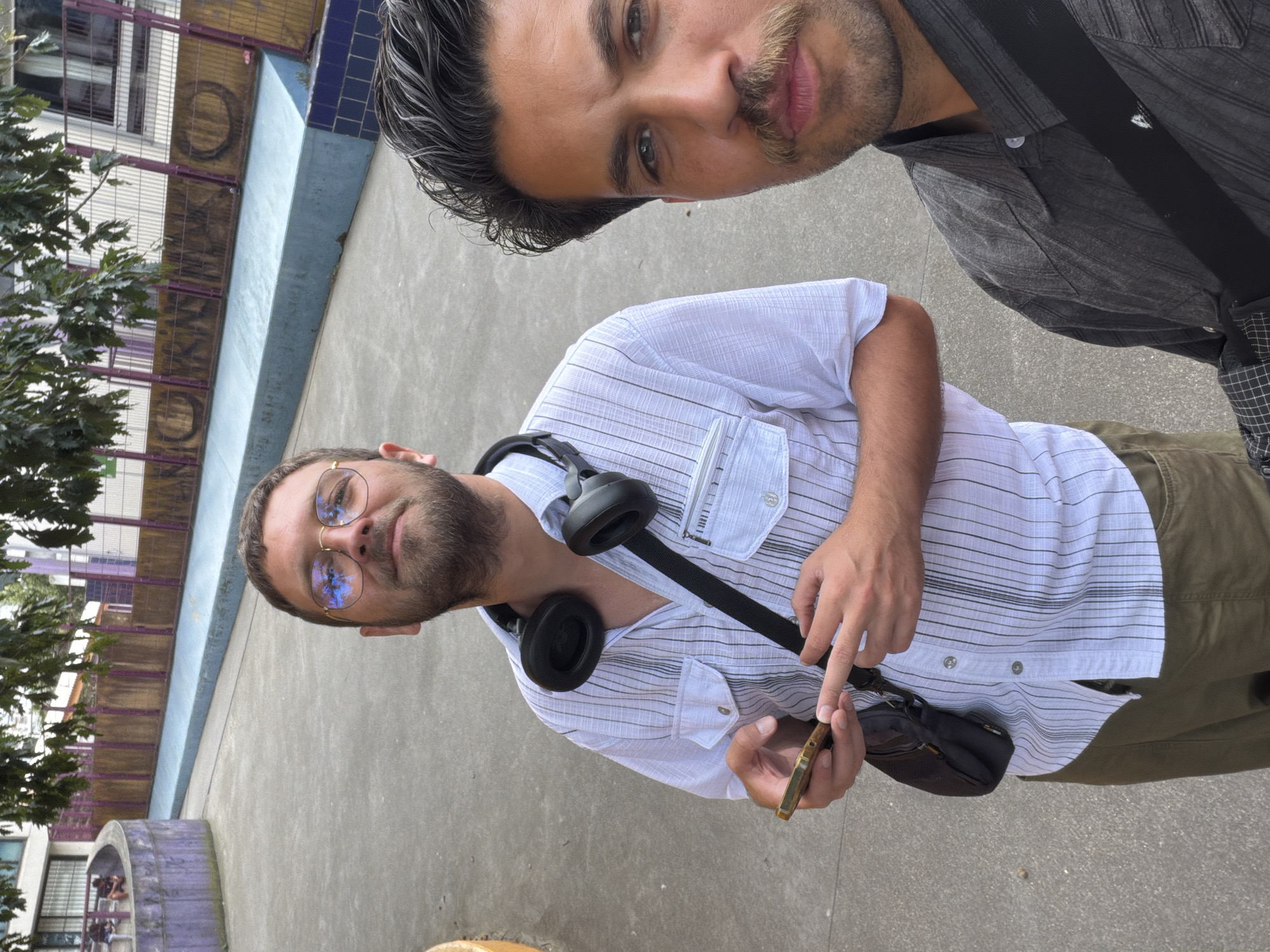 This media file shows two men in an outdoor setting. One man is closer to the camera, wearing a white striped shirt and khaki pants. He has headphones around his neck and is holding a smartphone in his right hand, appearing to interact with it. He is also wearing glasses that reflect blue light. The second man is partially visible in the upper right of the frame, wearing a dark grey patterned shirt. He has a mustache and appears to be looking towards the camera.
The background includes a textured concrete ground, a building with purple and yellow accents, and some greenery from a tree on the left. The setting appears to be urban, possibly a public space. The lighting suggests it is daytime. No specific activity or event is evident, beyond the individuals being present and one person using a phone.
Visible text includes "IXNO" in large letters on the building facade. The context provided indicates the location is Ivry-sur-Seine, France.