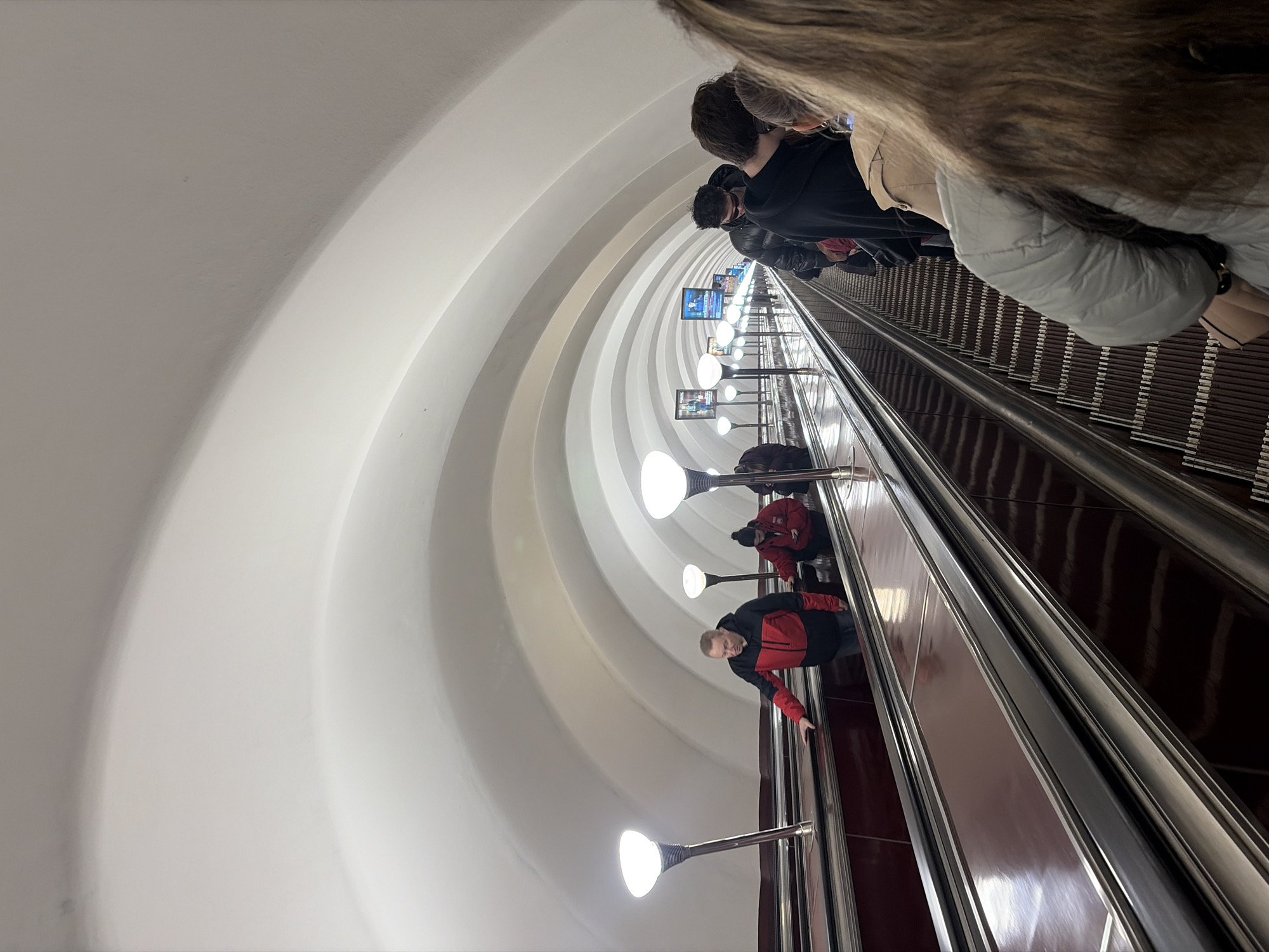 The image depicts a long, deep escalator descending into an underground metro station, characteristic of those found in Saint Petersburg, Russia. The perspective is from a slightly elevated position on the escalator, looking down its considerable length.
Several people are visible riding the escalator. In the immediate foreground, the back of a person with long, light brown hair is seen, wearing a light grey puffer jacket over a beige garment. Further down, other individuals in dark clothing are present. A man in a red and black jacket is notably positioned mid-frame, leaning against the silver handrail and looking towards the photographer. Another person in a red jacket is visible further down the escalator.
The escalator itself features dark brown steps and sturdy silver handrails. It is enclosed within a distinctive white, arched tunnel, characterized by multiple concentric rings that create a strong sense of depth and architectural grandeur. Along the descent, numerous circular, white-domed lights with silver bases are uniformly spaced, providing bright illumination to the tunnel. Further down the tunnel walls, several rectangular, illuminated screens display what appear to be advertisements or informational content; the text on them is too small to be legible. The scene depicts a typical underground transport environment, with passengers traveling between levels in an artificially lit setting. No specific time of day or weather is discernible.