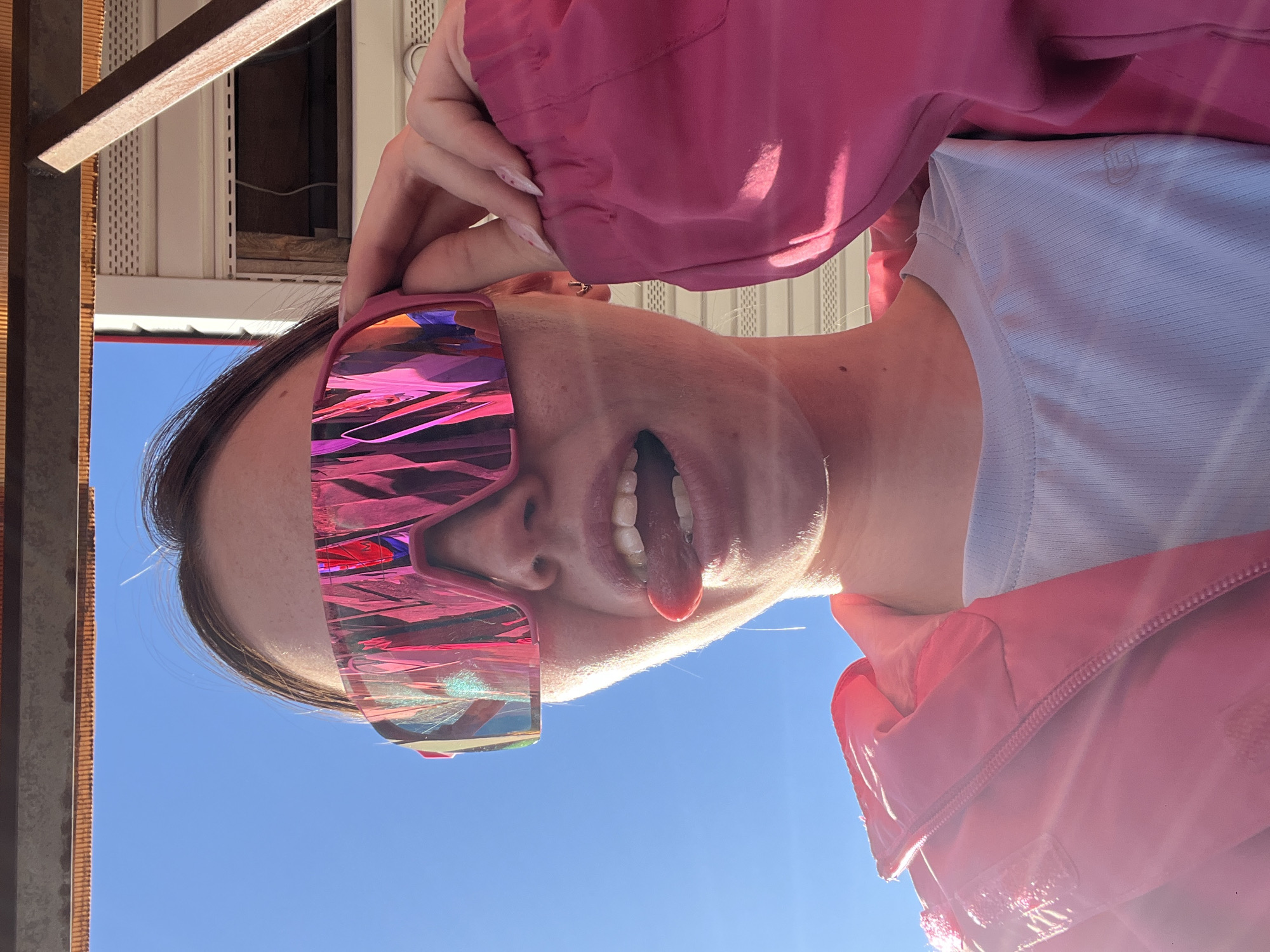 A young person is posing for a selfie outside in Gory, Russia. The subject is tilted at an angle, looking down at the camera with their tongue sticking out. They are wearing pink iridescent sunglasses and a pink jacket over a light blue shirt. The sky is blue and visible in the background. Part of a building structure is visible in the upper left corner. The setting appears to be a sunny day.
