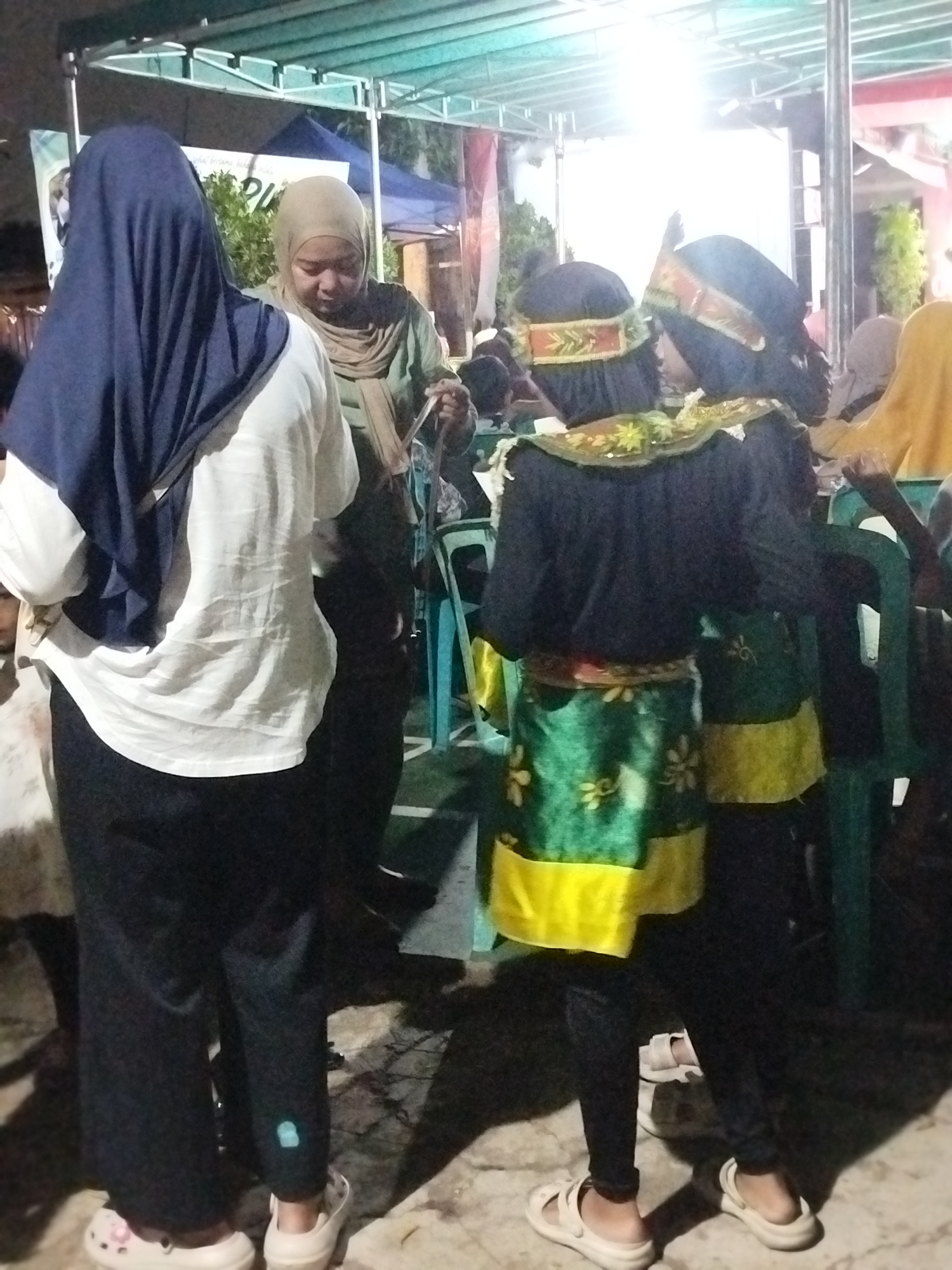 The image depicts an outdoor event, possibly a community gathering or cultural performance, in Depok, Indonesia. In the foreground, two individuals are dressed in traditional attire. One wears a black top with gold and green embellishments and a green skirt with floral patterns over black leggings and white sandals. The other, facing away from the camera, wears a similar outfit with a green skirt. To the left, a person in a dark blue hijab and white shirt stands with their back to the camera, also wearing black pants and light-colored footwear. Behind them, a person in a light brown hijab is visible. The background shows a tent-like structure with lighting, and other people are present, some seated on chairs. The ground appears to be paved. The overall lighting suggests it is evening or nighttime.