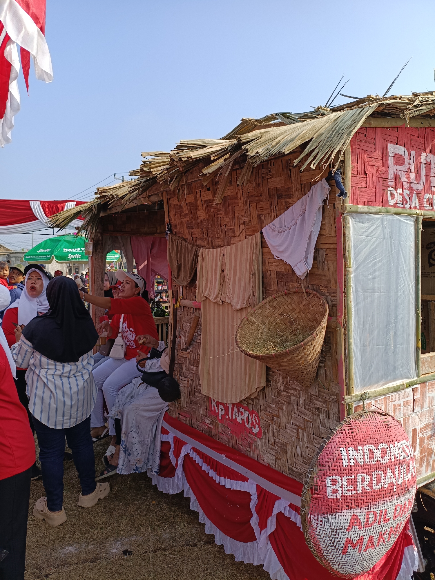 In the city of Cibodas, Indonesia, on a bright day, a temporary structure made of bamboo and dried palm leaves stands adorned with red and white fabric. People are gathered around, some interacting with each other, while others seem to be observing. The structure itself resembles a small booth or stall, with a woven bamboo exterior. Inside, there are various items visible, including clothing items hanging on the walls and a basket filled with straw. A large circular woven object, painted with red and white patterns and text, is attached to the side of the structure. The ground is covered in dry grass, indicating an outdoor setting. Red and white banners are also visible, suggesting a festive or celebratory occasion.