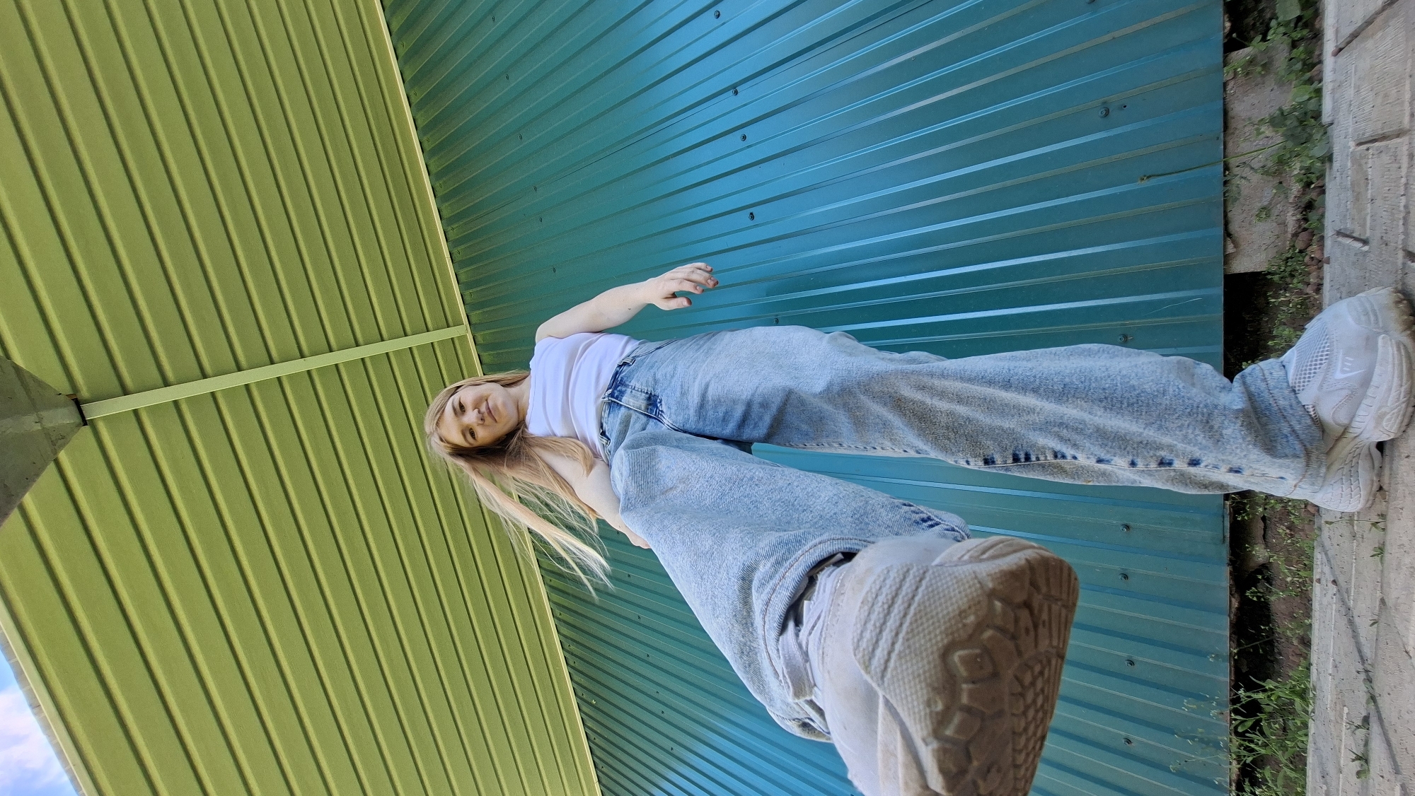 A young woman with long blonde hair is lying down and looking up at the camera. She is wearing a white t-shirt and light blue jeans, and white sneakers. The woman is positioned in front of a teal corrugated metal wall. To the left of the woman, there is a lime green corrugated metal structure, possibly a roof or awning, with a horizontal support beam. The image is taken from a low angle, looking up. Some greenery and a concrete or stone surface are visible to the right of the woman. The lighting suggests it is daytime, with natural light illuminating the scene. The overall impression is casual and relaxed. There is no visible text in the image. The location is specified as Bolshoy Bukor, Russia.