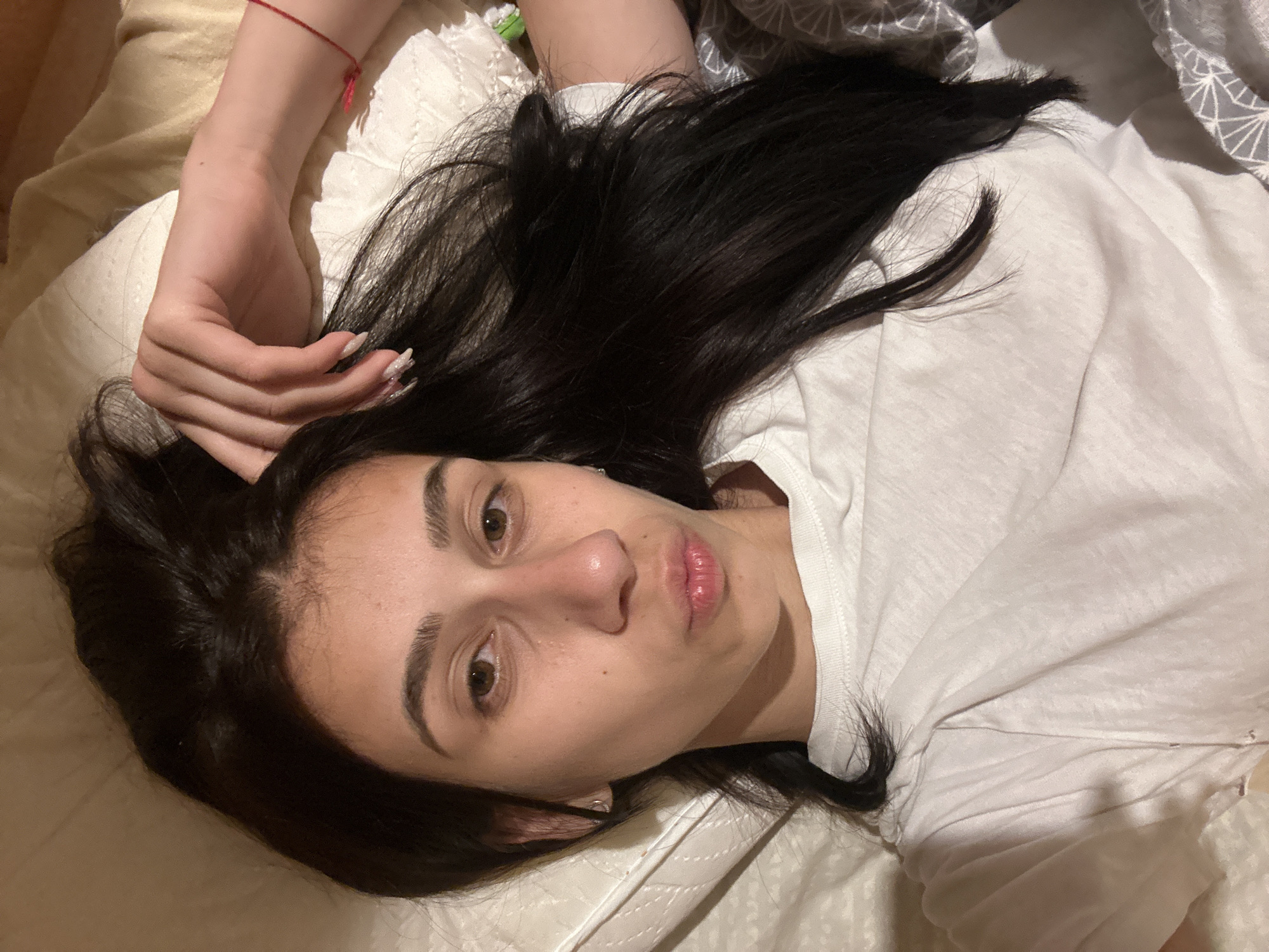 A young woman lies on a bed in what appears to be a domestic setting. She has dark hair and is wearing a white t-shirt. Her left arm is raised near her head, and she is looking directly at the camera. The background consists of bedding with varying textures and patterns. The image does not provide any information to determine the location within Plovdiv, Bulgaria. There are no actions, interactions, or other subjects visible in the frame.
