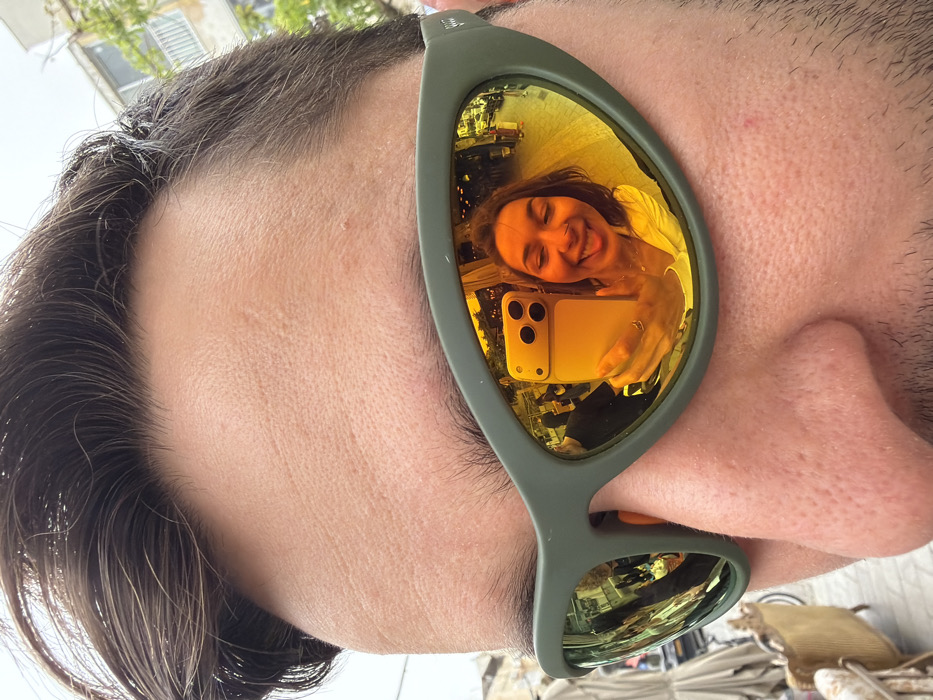 The image presents a close-up, first-person view of an individual's upper face, specifically focusing on the forehead, eyebrows, and the bridge of the nose. The subject has short, dark brown hair and fair skin. They are wearing olive green-framed, mirrored sunglasses.
The primary subject of interest is the reflection visible in the right sunglass lens (from the viewer's perspective, this is the left lens). This reflection shows a female individual taking a selfie. She has brown hair, is smiling with her tongue slightly extended, and holds a yellow smartphone (featuring a three-lens camera array) in her right hand, pointed towards herself. The reflection's warm, golden tint reveals an outdoor urban environment. Visible elements within the reflection include other indistinct people, the facades of buildings, and what appears to be street market stalls or outdoor cafe seating. An ornate, gold-painted chair, suggestive of antique furniture, is partially visible outside the reflection in the lower-left foreground of the main image, providing context to the reflected environment.
Based on the characteristic outdoor market ambiance, the presence of antique-style furniture, and older architectural elements, the scene is consistent with the Jaffa Flea Market (Shuk Hapishpeshim) area in Jaffa, Tel-Aviv, Israel.