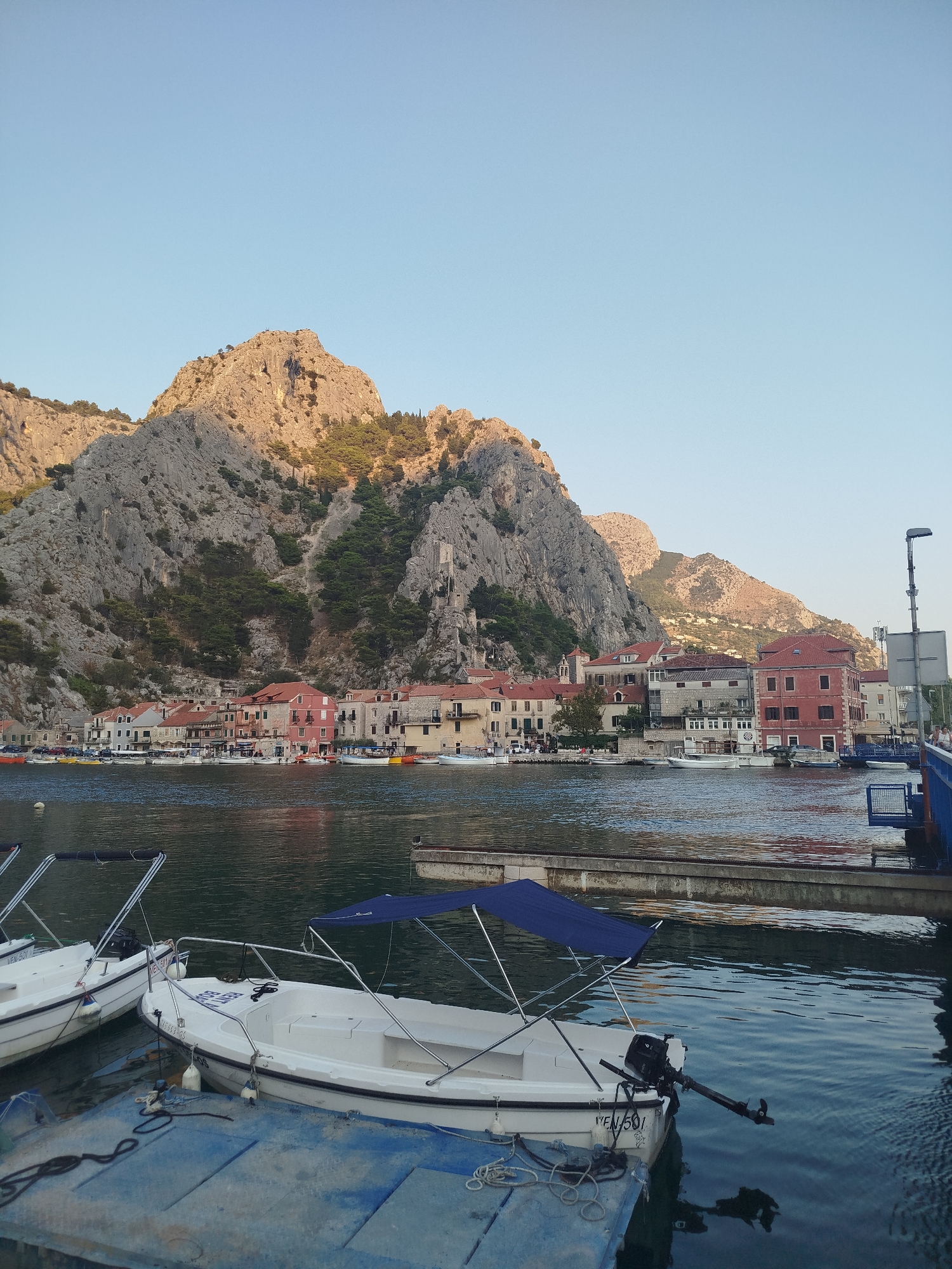 The image shows a view of the city of Omiš, Croatia, featuring a body of water with several boats docked in the foreground. The water is calm, reflecting the clear blue sky and the town's buildings. In the background, a range of steep, rocky mountains dominates the landscape, with the town nestled at their base. The architecture of Omiš is visible along the waterfront, characterized by buildings with red-tiled roofs. Two white motorboats are prominently featured in the foreground, tied to a blue dock. One boat has a blue canopy. The overall scene conveys a peaceful, coastal atmosphere.