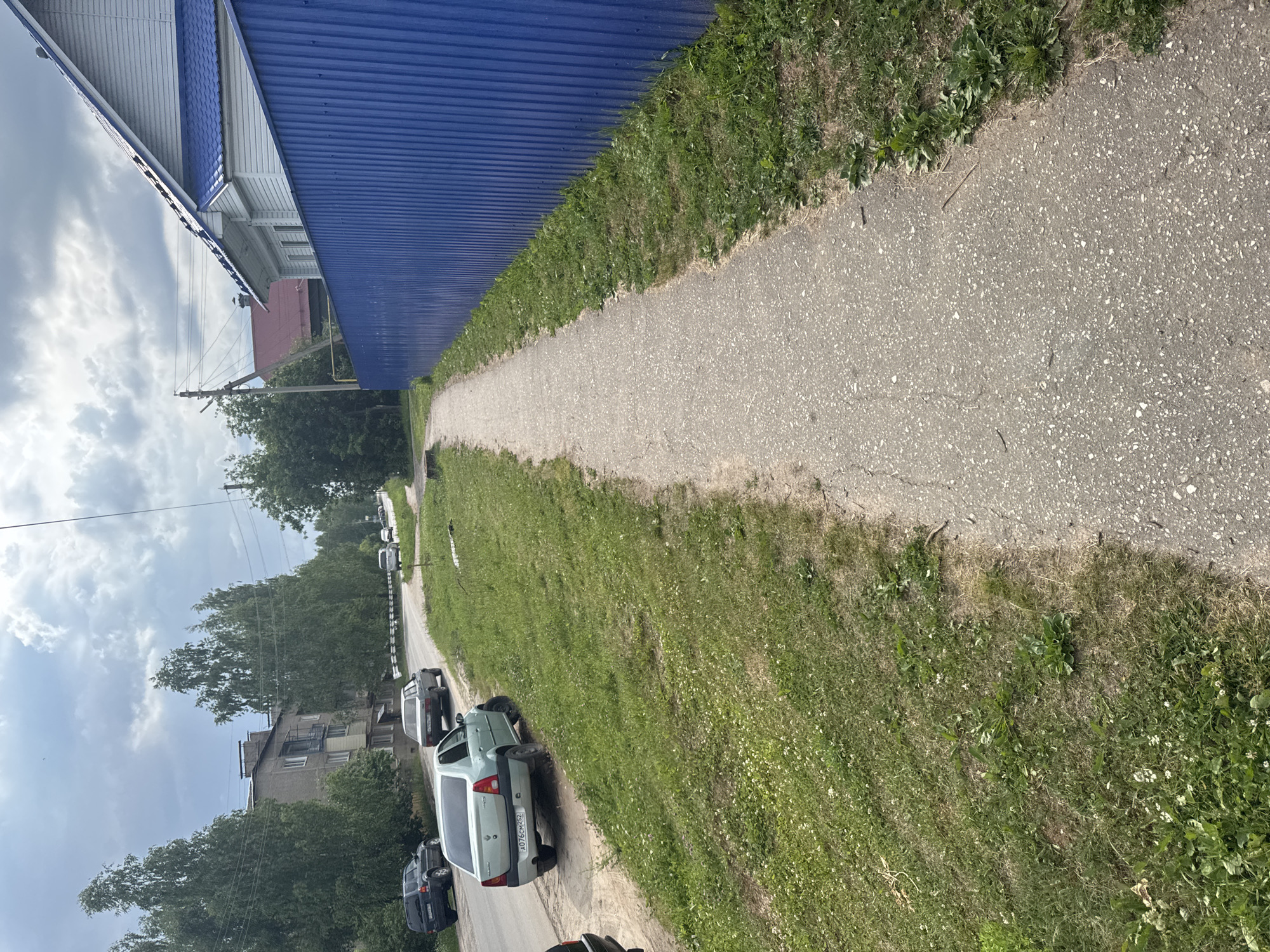A residential street in Krasnye Baki, Russia, features a paved sidewalk running alongside a bright blue corrugated metal fence. To the left of the sidewalk, a grassy verge is dotted with small white flowers, and beyond that, a paved road is visible with several cars parked or driving. One car, a light-colored sedan with license plate A076CM252, is parked on the grass verge. In the background, a multi-story apartment building and trees line the street. The sky is overcast with gray clouds.