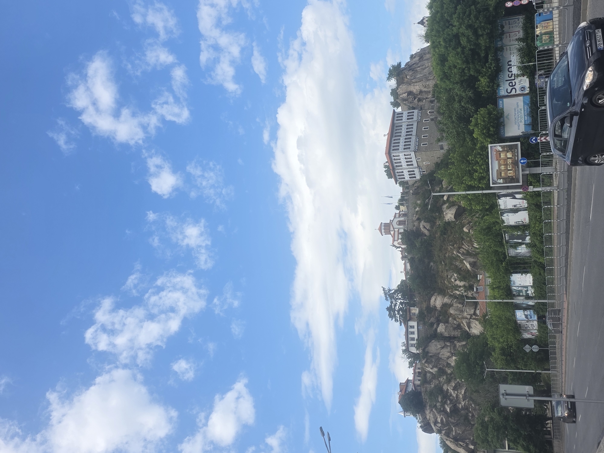 The image captures a view of a rock formation in Plovdiv, Bulgaria, featuring buildings perched atop it. The camera's perspective is from a ground level, looking slightly upwards, with a blue sky filled with scattered white clouds occupying the left and upper portions of the frame.
On the right side of the image, a road is visible, with a black car parked on it. A billboard display with advertising is situated along the road. The buildings on the rock formation vary in size and style, and are interspersed with greenery and trees. Streetlights and signage are also present along the roadside. The image is oriented vertically, which positions the sky to the left and the ground to the right.