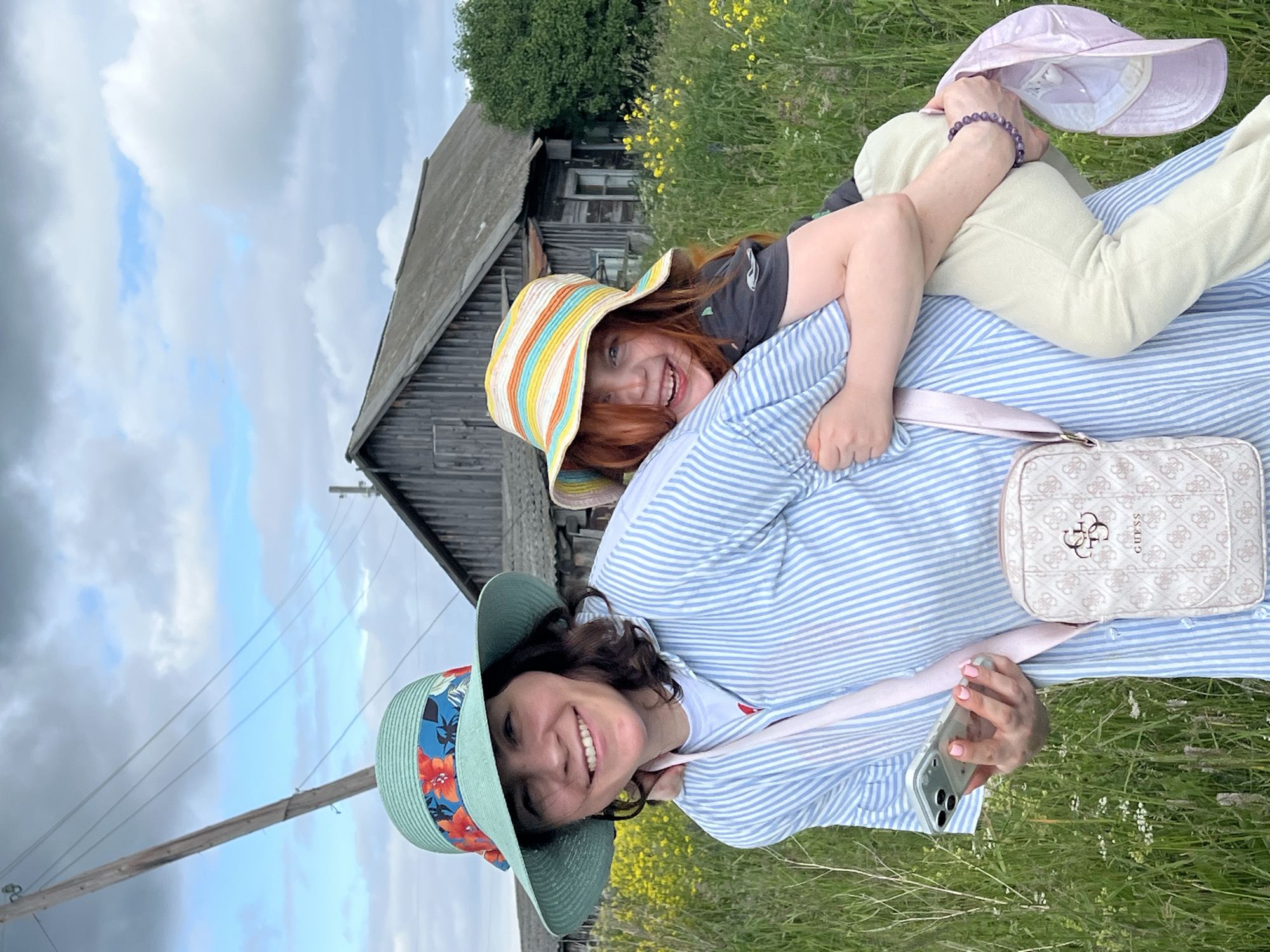 In the village of Panteleevka, Russia, two females are pictured in a grassy field under a cloudy sky. The female in the foreground is wearing a light blue and white striped shirt and a light green hat with a floral band. She is holding a smartphone in her left hand and has a crossbody bag with a "Guess" logo. The second female, who appears to be younger, is being carried on the back of the first female. She is wearing a striped hat and has reddish hair. The background shows a weathered wooden building, a utility pole with wires, and yellow wildflowers.