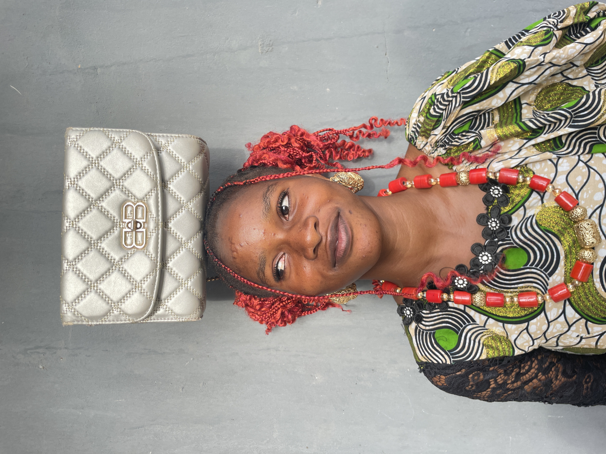 A young woman is lying on a light grey, possibly concrete, surface in what appears to be Badagry, Nigeria. She has dark skin and her head is turned, looking directly at the camera with a subtle, warm smile. Her hair is styled in bright red braids, some of which are wavy or curly.
She is adorned with striking accessories. A multi-strand necklace of prominent red cylindrical beads, interspersed with smaller gold and translucent beads, drapes around her neck. She also wears small, intricate gold earrings. Her attire includes a vibrant patterned top featuring bold designs in green, gold, black, and beige, with a black lace trim visible on the sleeve.
Adjacent to her head, on the surface, rests a metallic silver or light gold quilted handbag. The handbag features a prominent metallic gold clasp or logo on its flap, consisting of two stylized, interlocking 'B' letters. The overall impression is a vibrant and composed portrait.