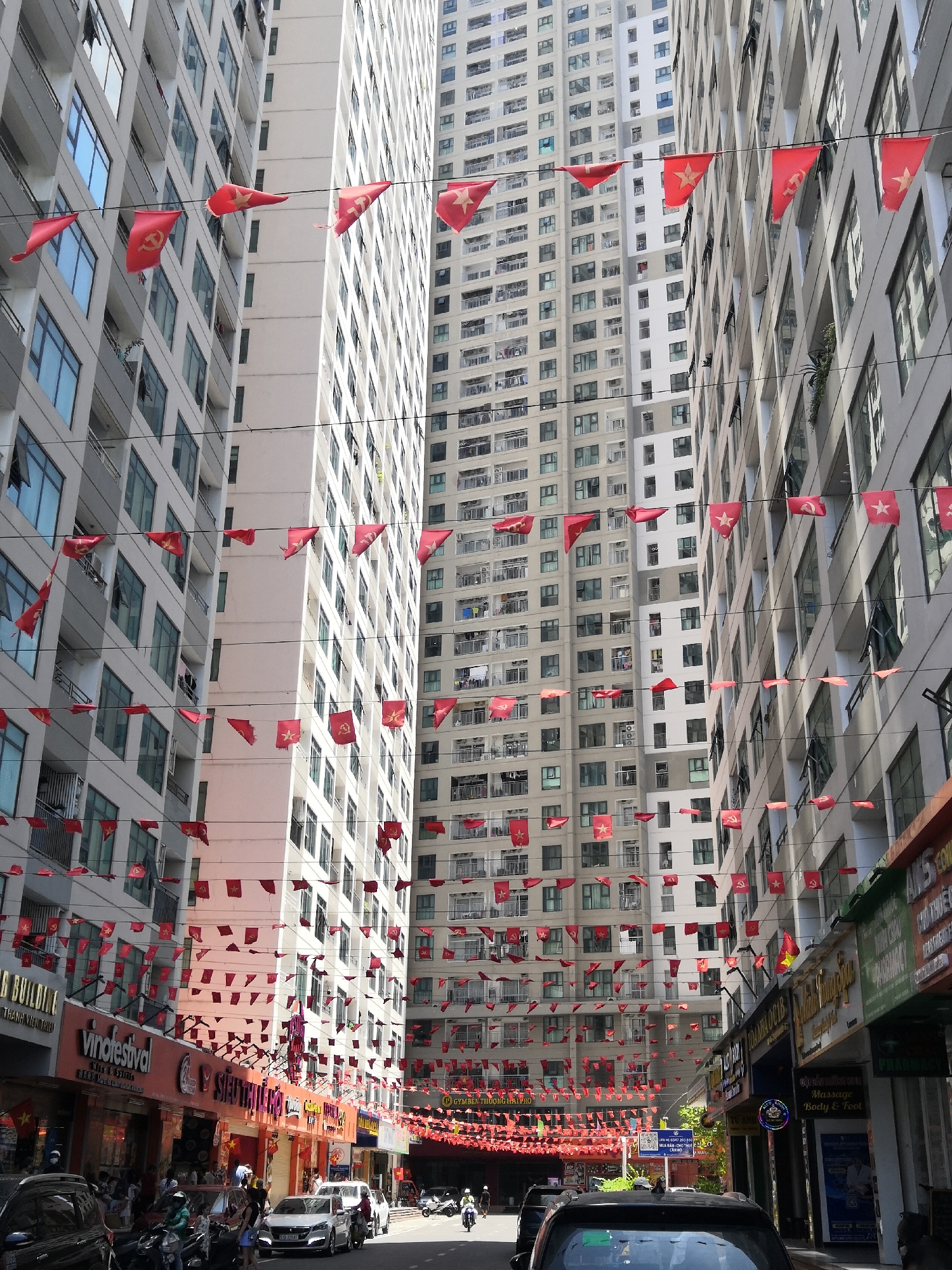 This image depicts a bustling urban street scene in Bac Nha Trang Ward, Vietnam, characterized by a dense display of red pennant flags strung overhead. High-rise apartment buildings, predominantly light-colored with numerous windows and balconies, line both sides of the street, forming a narrow corridor. Many windows are open, and laundry is visible on some balconies. Hundreds of small red pennant flags, featuring either a single yellow five-pointed star (symbolizing the Vietnamese national flag) or a yellow hammer and sickle (symbolizing the Communist Party of Vietnam), are densely strung on multiple wires crisscrossing the street at various heights, creating an overhead canopy. The street level features active commercial establishments with various signs, including "vinofestival" (wine & spirits), "SIÊU THỊ" (supermarket), "PHARMACY", and "Massage Body & Foot", among others in Vietnamese script. Multiple vehicles are present, including cars (a black SUV in the immediate foreground, a silver sedan further back) and numerous motorcycles and scooters, both parked and in motion. Several individuals are visible walking on the sidewalks and riding motorcycles, some wearing helmets. The scene is illuminated by bright daylight, casting visible shadows.