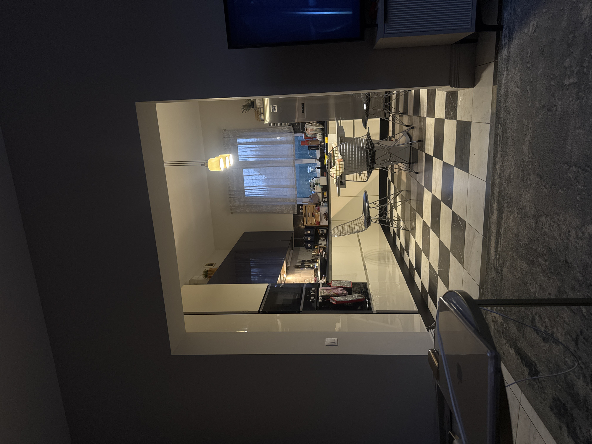 The image shows a modern kitchen with a checkered floor, seen through a doorway from an adjacent room. The kitchen features white and dark grey cabinets, a cooking range with food on it, and a window with sheer curtains. Chrome chairs are visible at a counter. The location is not discernible from the image. No people are visible.