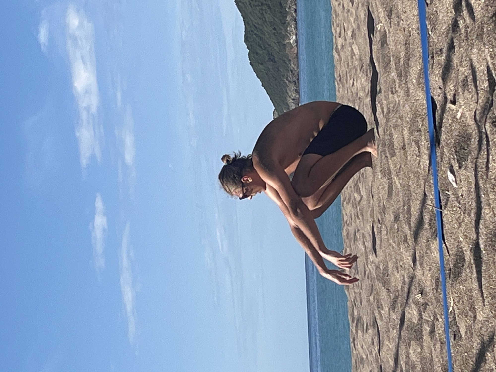 A man wearing dark swim trunks stands on a beach near Bečići, Montenegro. He is crouched, his arms extended downwards as if preparing to jump or dive. He is facing the ocean and wears dark sunglasses. The beach is composed of dark sand and is marked by a blue line. Behind the beach is a body of water, and beyond that, a green mountainous area. The sky above is blue with scattered white clouds.