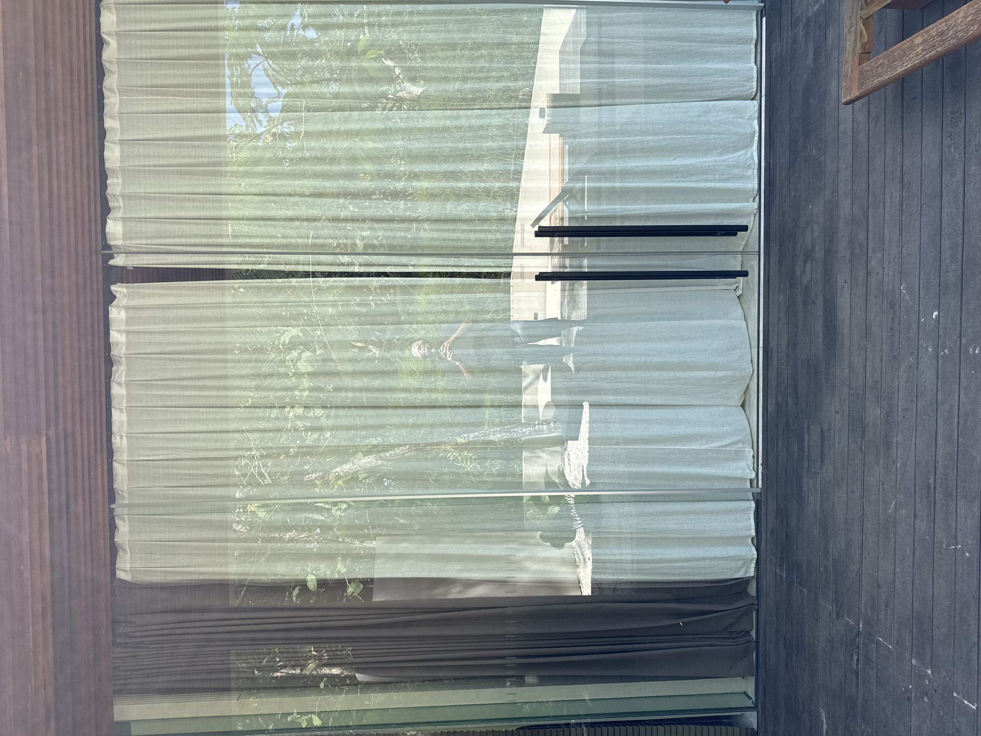 The image shows a window with layered white and dark curtains, with a blurred reflection of trees and a person visible through the window. Outside, part of a wooden deck and possibly a chair leg are visible. The photo seems to have been taken indoors. There's no explicit indication of the location being in Kuta Selatan, Indonesia, beyond what may be implied by the presence of tropical trees.