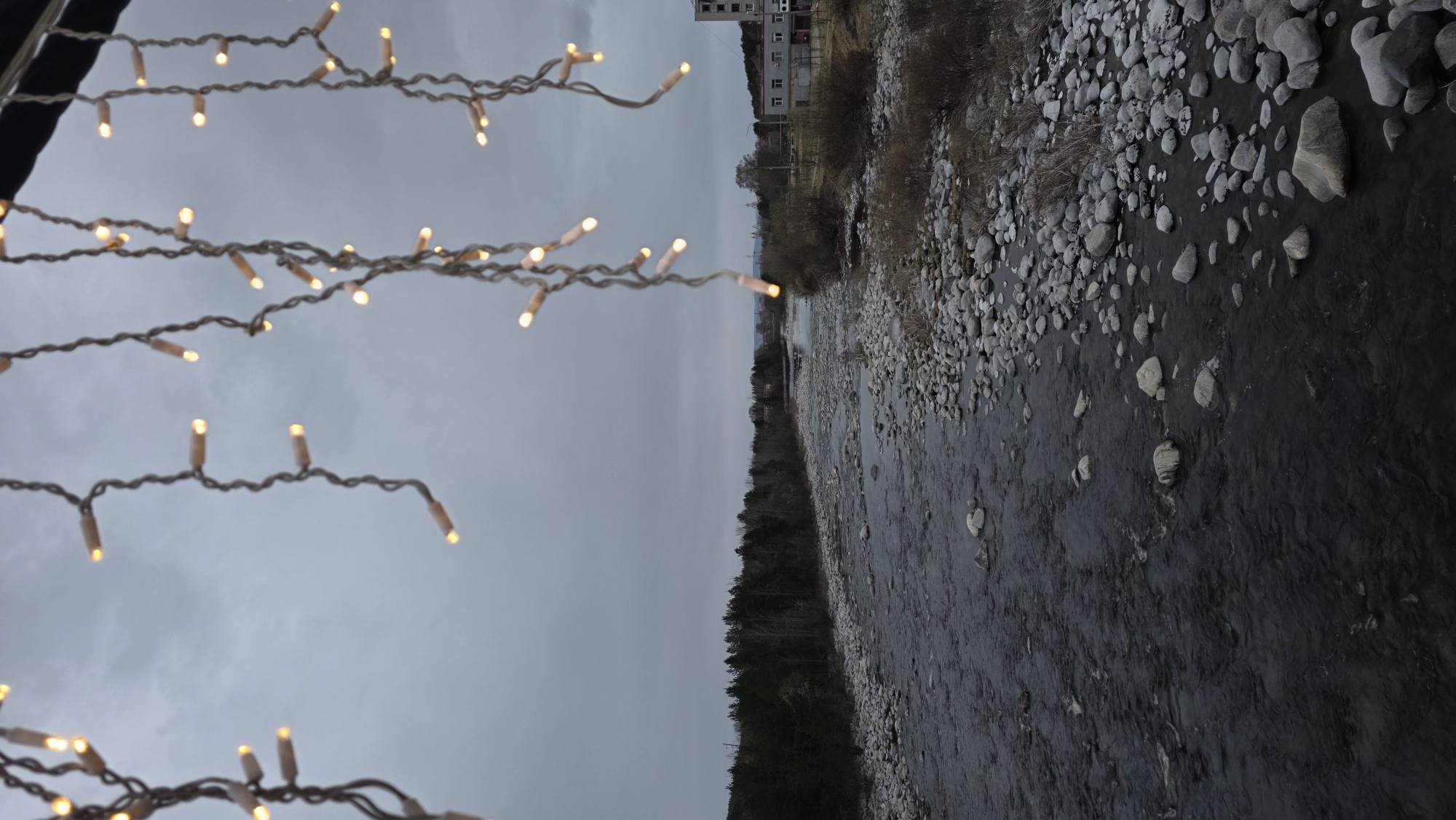 The image shows a river flowing through Kandalaksha, Russia. The riverbed is rocky, with stones of varying sizes visible along the banks and within the water. The water is dark, suggesting depth or low light conditions. The surrounding landscape includes a dense forest on one side of the river and a mix of vegetation and buildings on the other. The sky is overcast and gray, contributing to a somber atmosphere. A string of decorative lights is draped across the foreground, possibly indicating a bridge or an event space near the river.