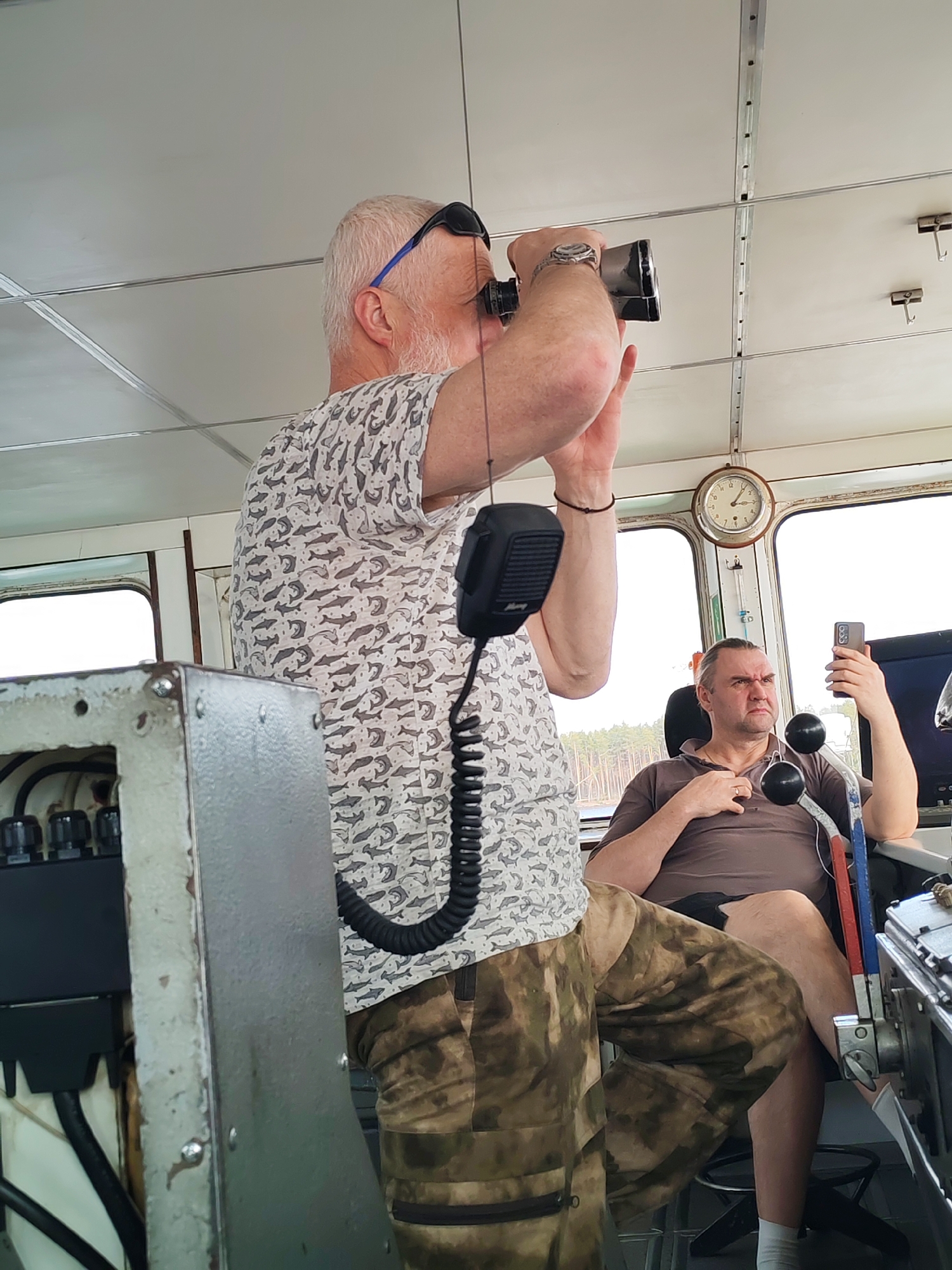 A medium shot captures two men inside a boat cabin. The first man, likely in his 60s, stands while looking through binoculars. He is wearing a light-colored shirt patterned with gray fish, camouflage pants, and sunglasses perched on his head. A microphone hangs from his shirt. The second man, appearing to be in his 40s, is seated and holding a smartphone, looking at the camera with a slightly furrowed brow. He is wearing a brown polo shirt and black shorts. In the background, there is a view of trees and water through the boat's windows. Other objects in the cabin include a clock, navigational equipment, and various switches and levers. The setting appears to be an indoor space aboard a boat, but the specific location is not identifiable from the visual evidence.