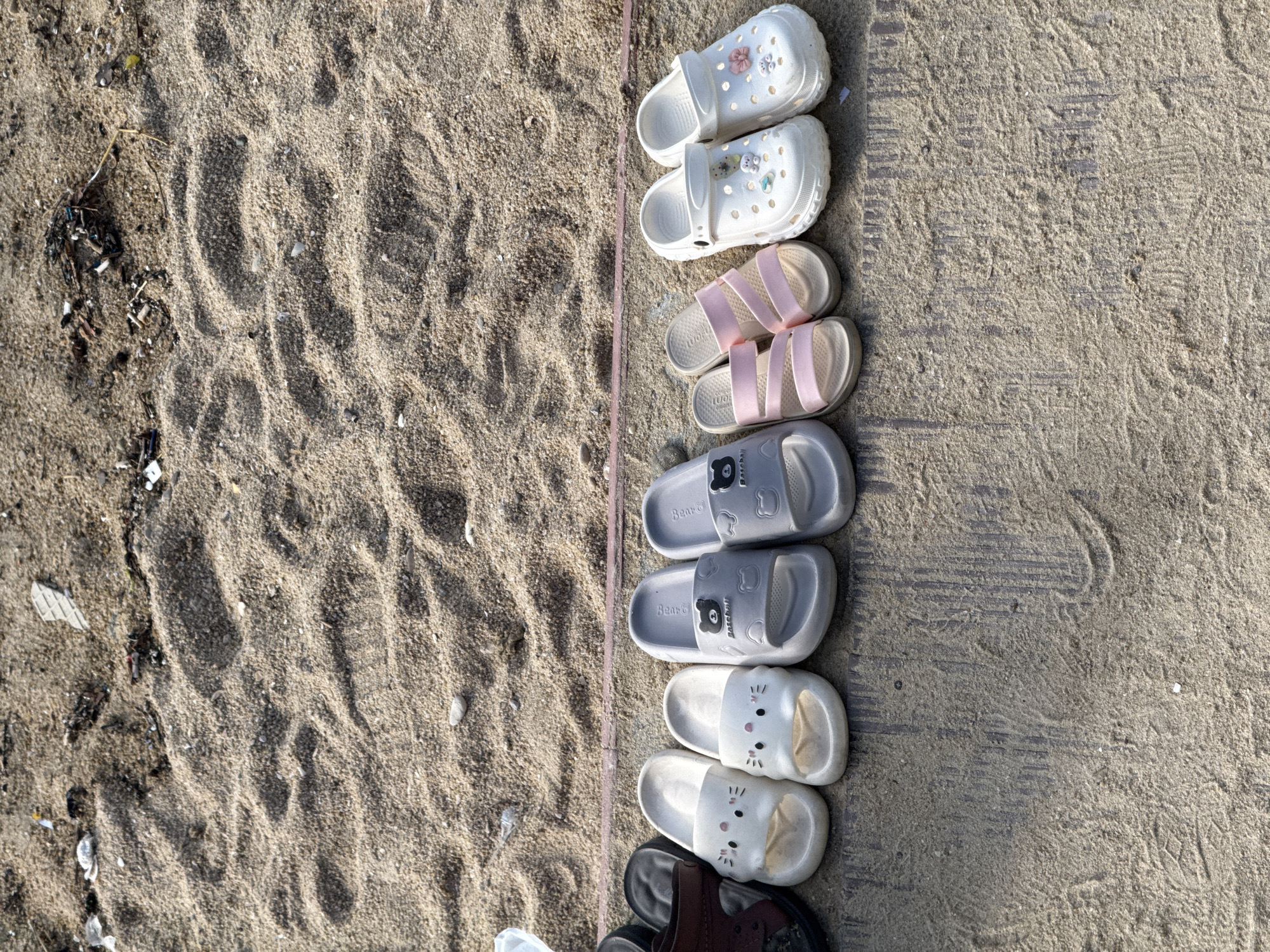 A row of ten sandals and flip-flops are arranged on a concrete surface next to a sandy area. The footwear includes two pairs of white sandals with cartoon cat faces, two pairs of grey slides with bear faces, a pair of pink strap sandals, and two pairs of white clogs decorated with small floral and bear charms. Behind the footwear, the sandy surface shows footprints and scattered debris. The concrete appears weathered and has a parallel line pattern. The image is taken from a high angle, looking down. The overall scene suggests an outdoor location, potentially a beach or a public area in Longgang District, China, where footwear has been removed.