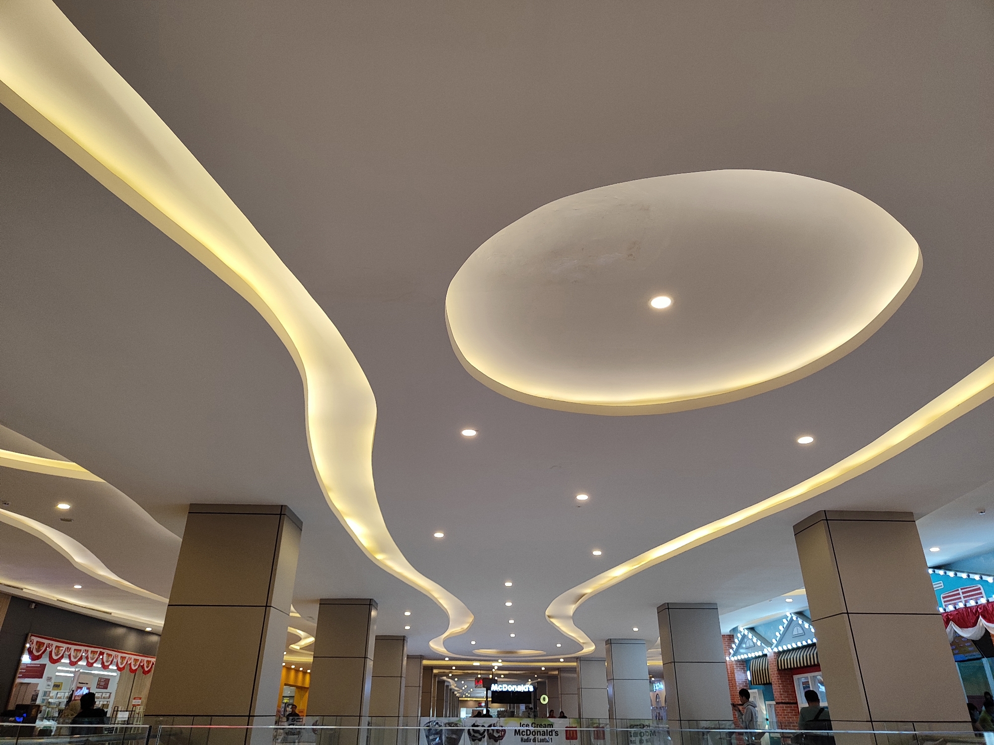 The image depicts the interior of a modern shopping mall, with a focus on the illuminated ceiling design. The ceiling features flowing, curved elements with integrated warm yellow lighting, creating a dynamic and contemporary aesthetic. Several circular recessed lights are also visible. Large, square pillars with a neutral, matte finish support the ceiling. In the background, glimpses of various retail establishments can be seen, including a McDonald's restaurant with its distinctive signage. People are present in the lower portion of the image, some browsing shops and others standing near the railing of an upper level, suggesting a typical mall environment. The overall impression is one of a well-lit and spacious commercial interior. The location is identified as Sempusari, Indonesia.