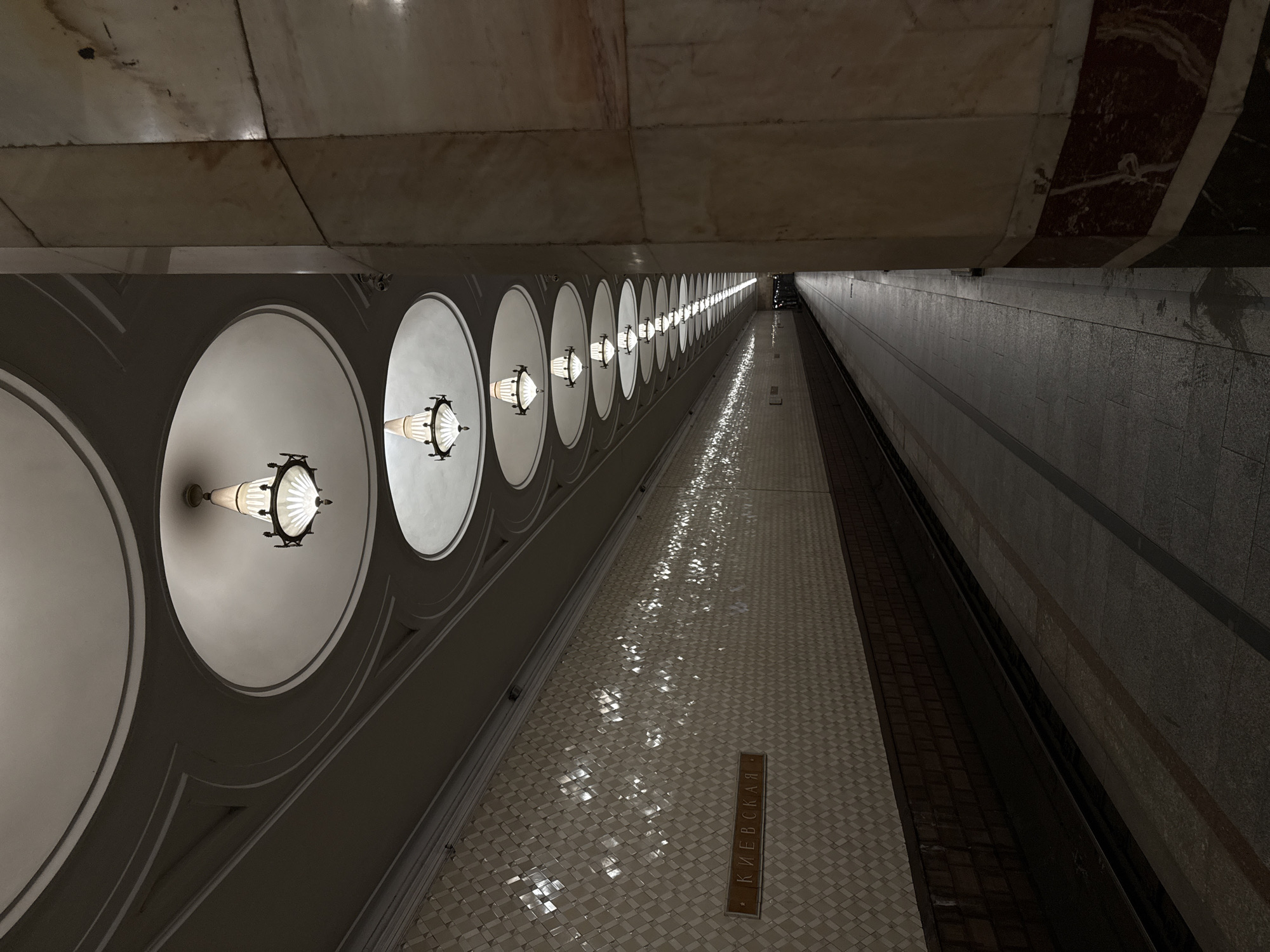 This image shows the platform of the Kievskaya metro station in Moscow, Russia. The platform features a repetitive design of large circular niches with ornate lamps. The floor is tiled in a mosaic pattern, and a sign reading "Kievskaya" is visible. The adjacent wall is dark and textured. No people are visible in the image.