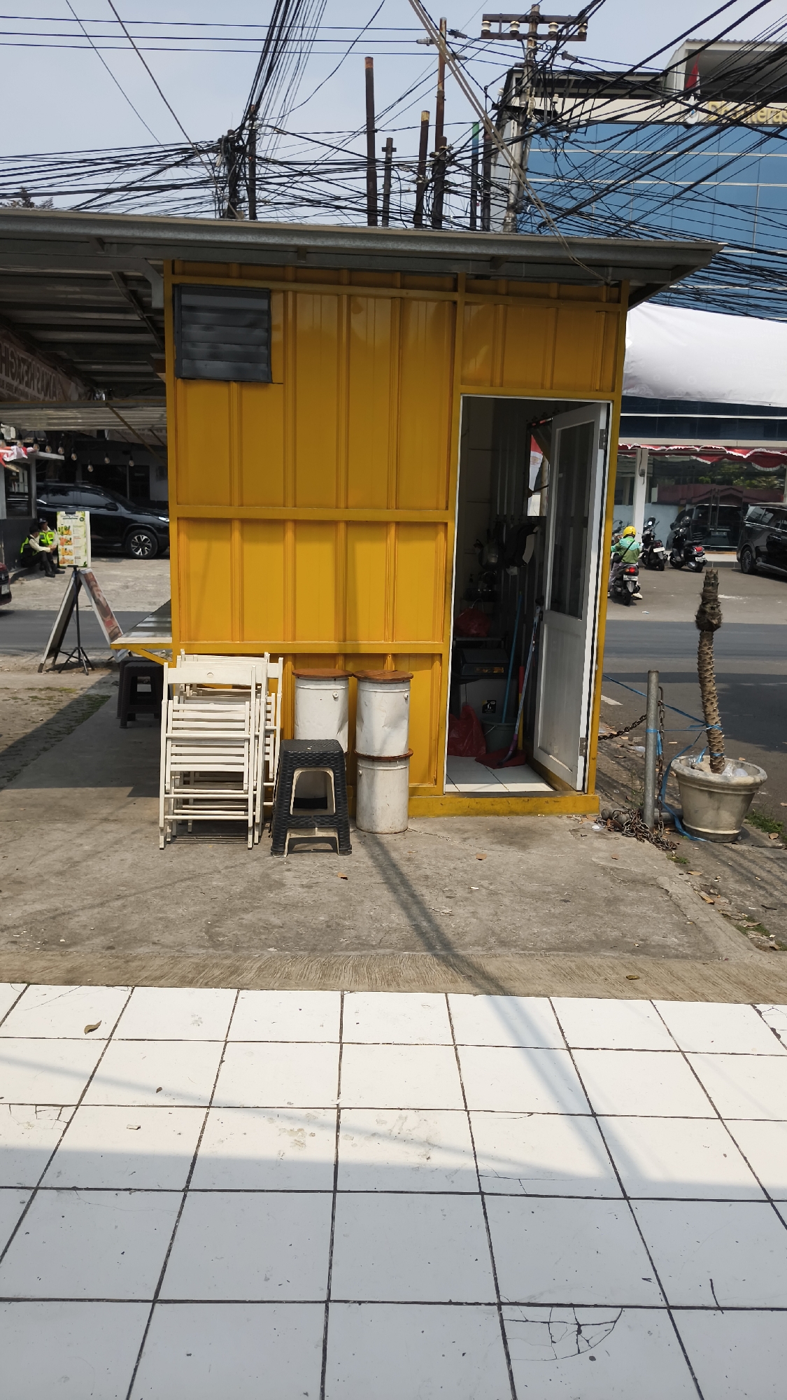 A yellow metal kiosk stands on a concrete sidewalk in South Jakarta, Indonesia, under a canopy. To the left of the kiosk, a stack of white folding chairs and a black stool are placed. Two large white cylindrical containers are stacked one on top of the other in front of the kiosk, with another identical container next to them. The kiosk has an open door revealing tools and cleaning supplies inside. A thin, dry palm tree in a pot is situated to the right of the kiosk, secured by a metal pole and chain. A street with parked motorcycles and cars is visible in the background, along with a dense network of overhead electrical wires and poles. The sky is overcast.
