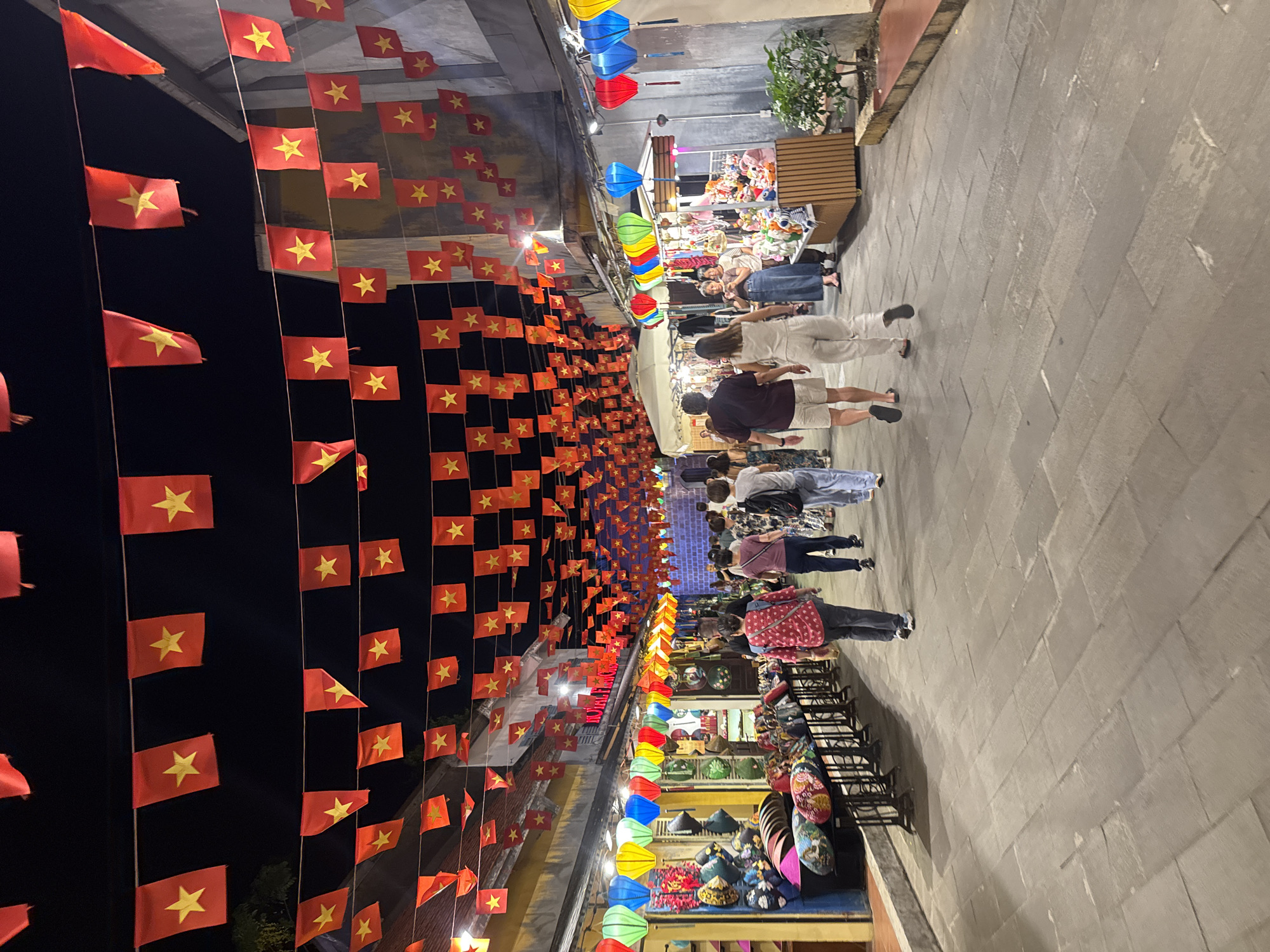 The image depicts a street scene at night in Hội An Ward, Vietnam. Above, strings of Vietnamese flags hang in rows across the street. Colorful lanterns adorn the sides of the street. People stroll down the sidewalk, looking at stalls with various wares. The walkway is paved with light gray pavers.