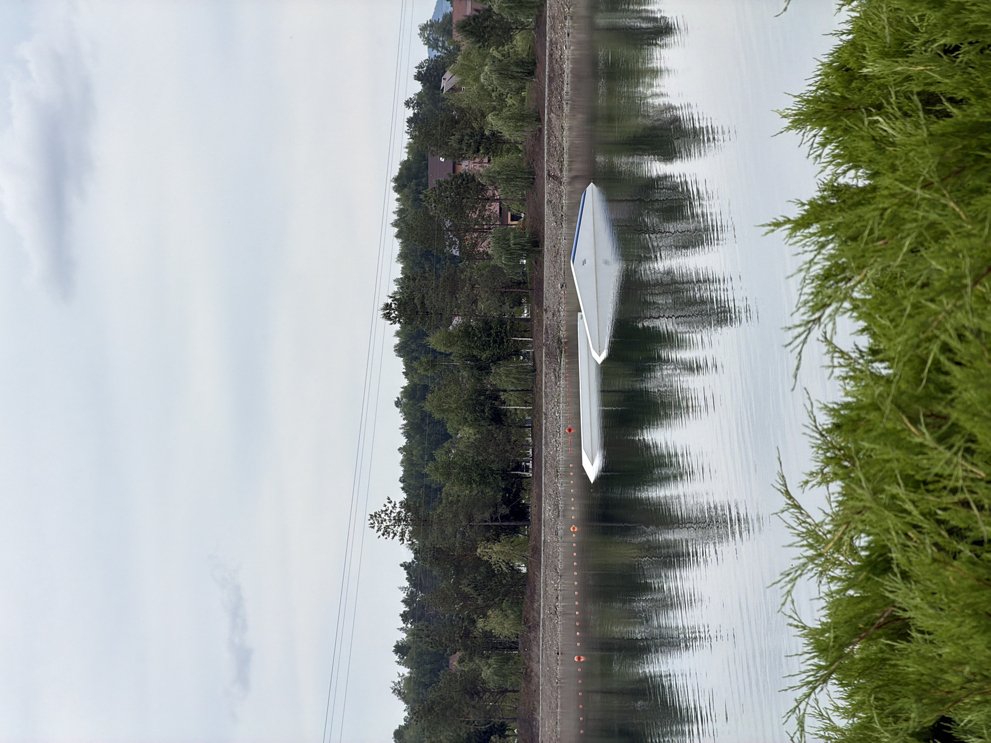 The image depicts a wake park located on a lake in the city of Озерное (майминский), Russia. In the foreground, lush green bushes line the bottom of the frame, partially obscuring the view. The lake itself is calm, with a smooth surface reflecting the overcast sky and the surrounding environment. Several orange buoys are visible in the water, marking a course or area.
Dominating the mid-ground of the lake are two white wakeboard ramps. One ramp is partially submerged, with its angled surface jutting out of the water, while the other is more fully visible, also angled. These structures suggest active use of the lake for water sports.
In the background, a treeline of various green trees forms a dense backdrop, with some houses and buildings nestled among the foliage, indicating a populated area bordering the park. Power lines are strung across the upper portion of the image, crossing the sky. The sky is filled with light clouds, suggesting a cool, possibly humid day. The overall scene is serene and suggests a recreational outdoor setting.