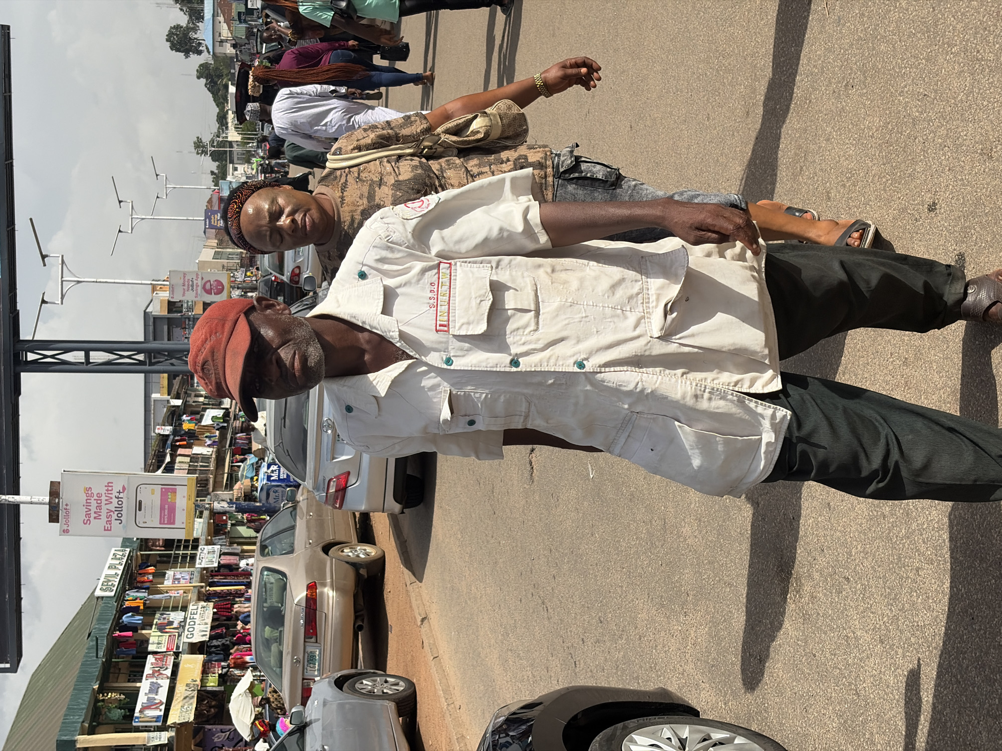 The image depicts a street scene in Jos, Nigeria, likely in an area with commercial activity. In the foreground, a Black man wearing a red cap and a light-colored, long-sleeved shirt with pockets and green buttons walks towards the viewer. The shirt has a patch with red and blue lettering that reads "S.S.D.I.O" and "INURTW". He is also wearing dark pants. To his right and slightly behind him, a Black woman with a patterned headband and a camouflage-patterned shirt is visible, carrying a beige bag.
The background shows a street lined with shops and stalls displaying various goods, including clothing. Several cars are parked or driving on the street. A prominent advertising billboard for "Jolloft+" is visible on a pole. The sky is overcast. The overall impression is of a busy, everyday urban environment in Jos.