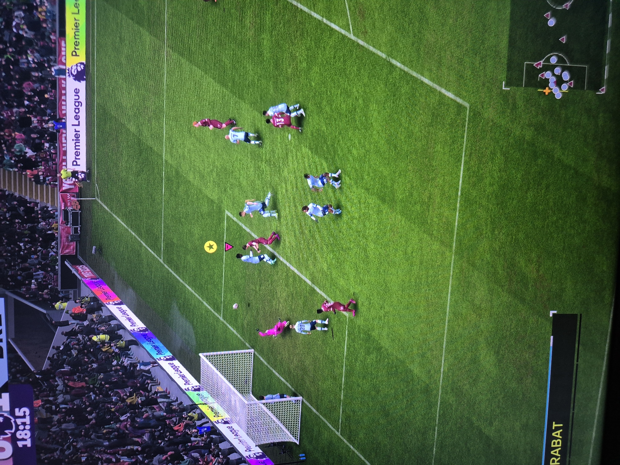 The image shows a soccer match being played on a grass field. The game is being broadcast on a screen, as indicated by the score overlay at the top left which reads "18:15". There are many players on the field, wearing different colored uniforms. Some players are actively engaged in the game, while others are positioned around the field. A goalkeeper in a pink uniform is diving to save a ball near the goal. Spectators are visible in the stands behind the field, suggesting this is a live event or a recording of one. The stadium features advertising boards along the sidelines, displaying "Premier League" logos and messaging. A mini-map or tactical view is visible in the bottom right corner, showing player positions. The overall environment is a sports stadium.
