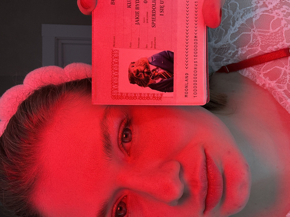 A person's face dominates the frame, intensely lit in red, with eyes open and lips slightly parted. A document, possibly a passport, obscures part of the forehead and displays text in a foreign language (possibly Polish). A smaller photograph on the document features a figure in a suit. Text on the document includes "MOONLAND" and a series of arrows followed by the characters "100000000RUS1000000M". A white, lacy fabric is visible in the upper right corner. The overall impression is surreal and intriguing, due to the red lighting and unusual composition.
