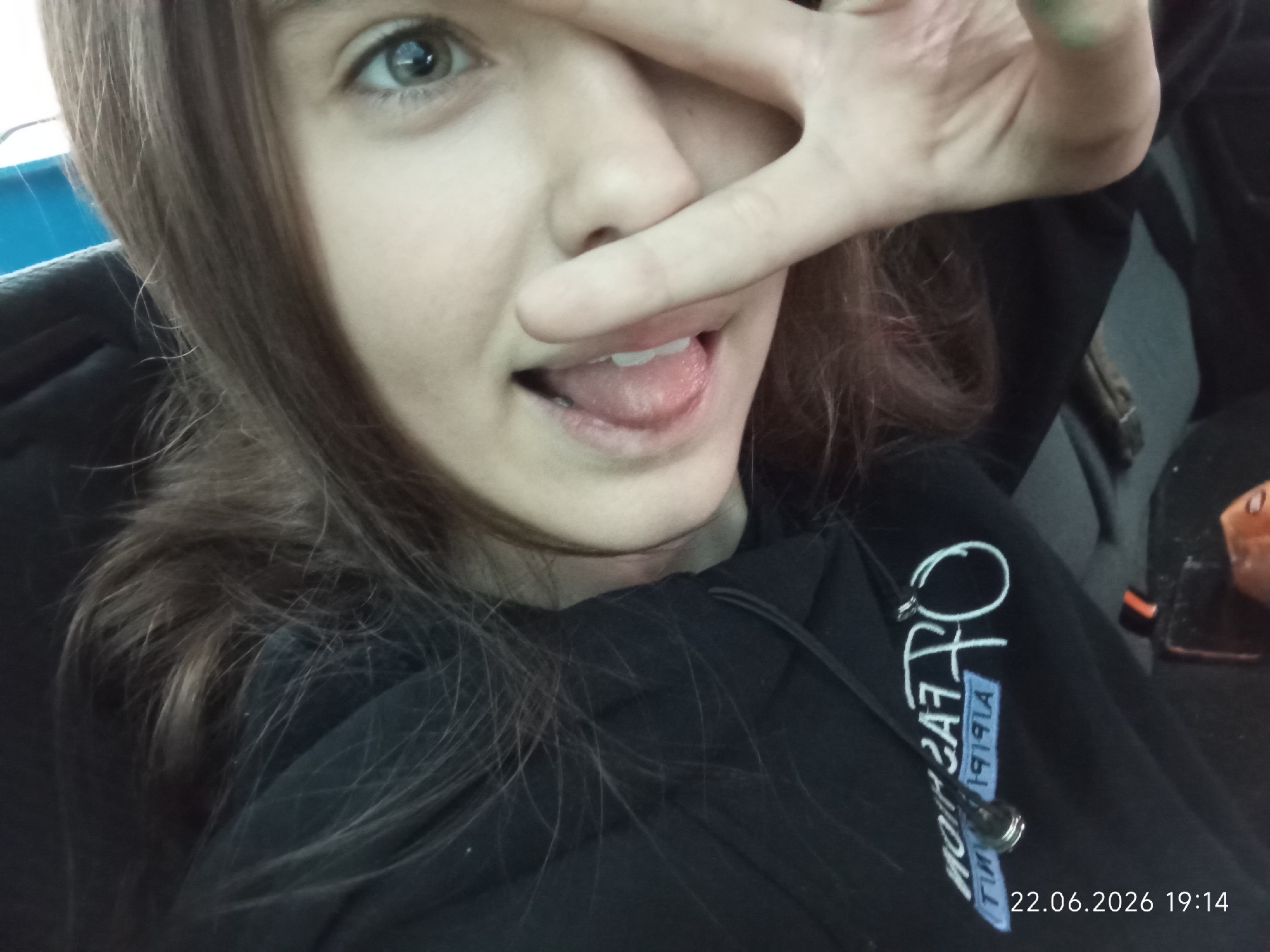 A close-up, selfie-style shot shows a young person with light brown hair and green eyes. They are wearing a black hooded sweatshirt and are making a peace sign over their eye. Their tongue is slightly sticking out. A date stamp in the lower right corner reads "22.06.2026 19:14". The background appears to be the interior of a vehicle, with a grey seat visible. There is no indication of the location being Sarytogay, Kazakhstan.