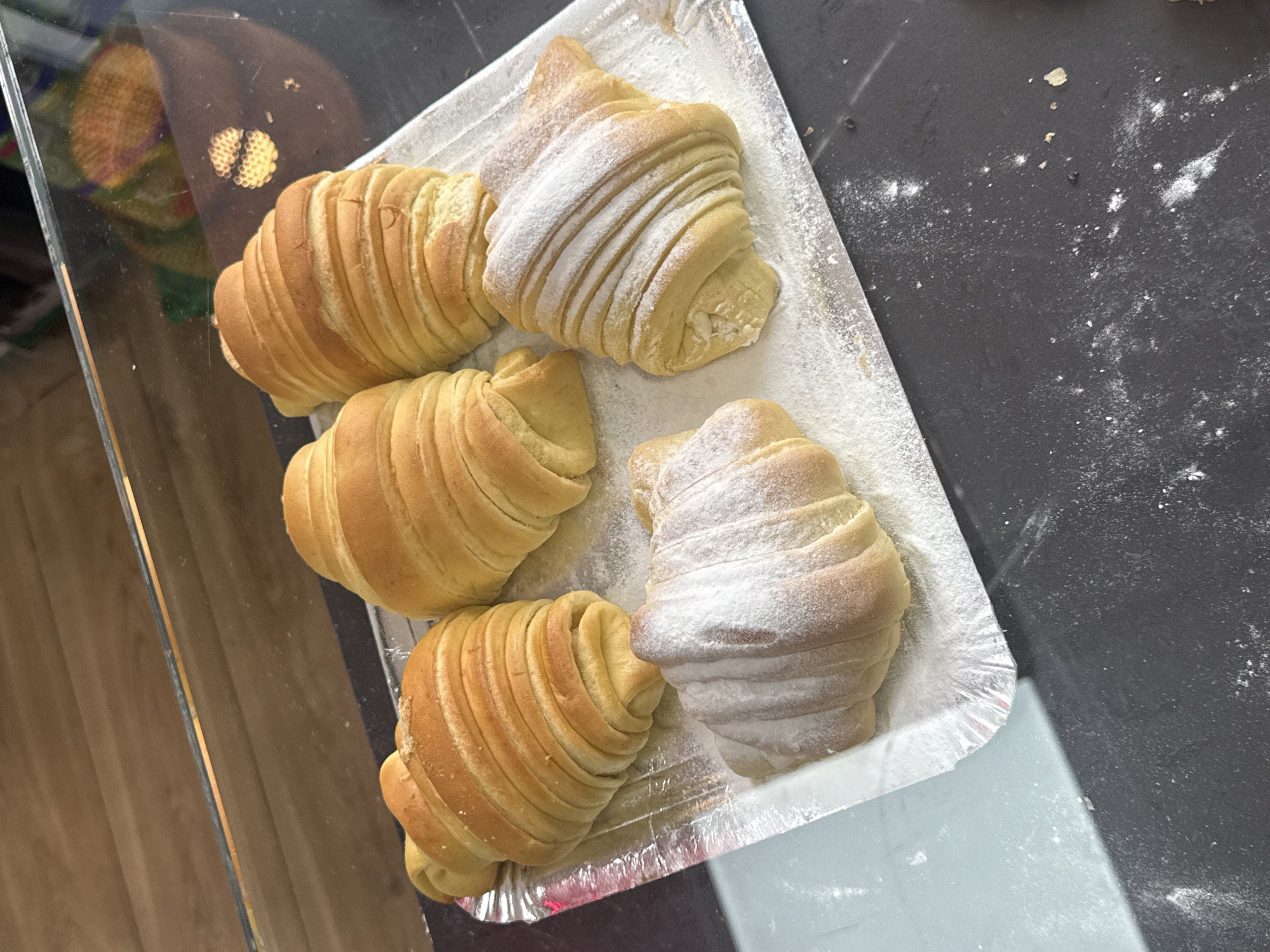 The image captures five freshly baked pastries, resembling croissants or crescent rolls, arranged on a rectangular, silver-colored, possibly foil-lined tray. Four of the pastries are golden brown, featuring distinct, parallel ridges that indicate a layered dough structure. The fifth pastry, positioned towards the top center of the tray, is lighter in color and generously coated with white powdered sugar, which also highlights its ridged surface. Another pastry, at the bottom right, also shows a lighter dusting of powdered sugar.
The tray is situated on a dark, flat surface, possibly a countertop, which has scattered white residue, likely flour or powdered sugar. To the left of the tray, a clear, reflective surface, possibly glass, creates a reflection of a blurred, patterned object, suggesting a display case or a retail setting. No people are visible, and the scene implies an indoor commercial environment such as a bakery or cafe. There is no discernible text within the image.