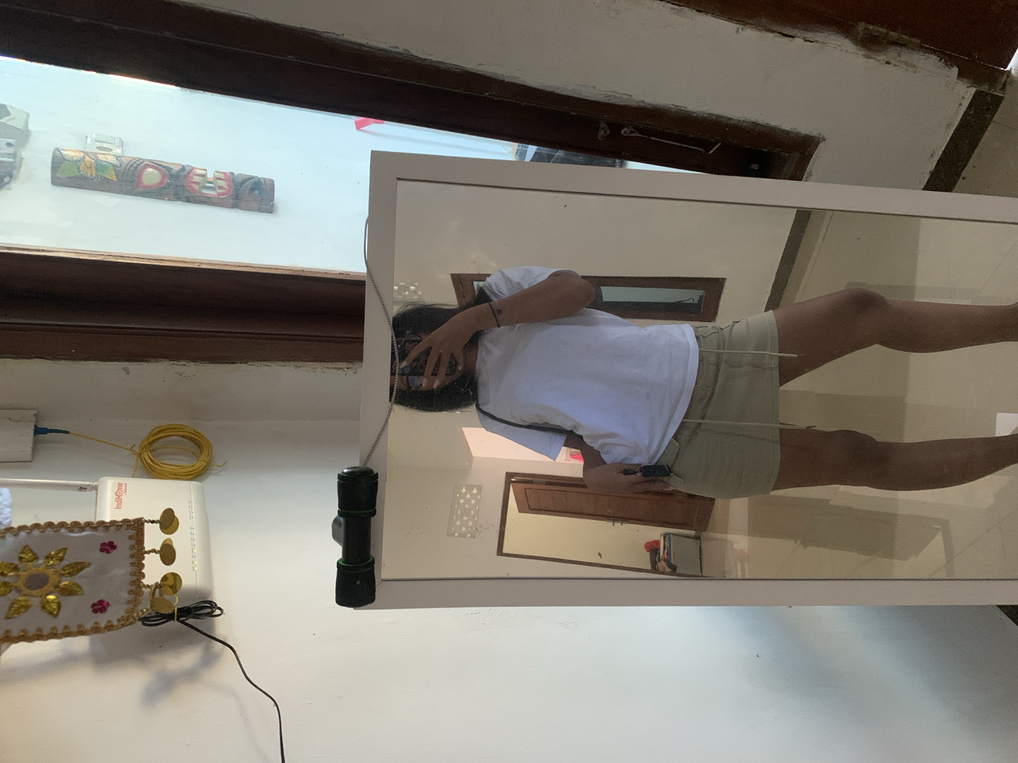The image depicts a person standing in front of a full-length mirror, taking a selfie. The person is wearing a white t-shirt and a khaki-colored skirt. The mirror reflects a room with tiled floors and white walls. To the left of the mirror, a white router is mounted on the wall, with yellow and black wires connected to it. A decorative fabric banner with gold embellishments hangs above the router. A black flashlight-like object is attached to the top of the mirror frame. In the reflection, a doorway is visible, with another room beyond it. The scene appears to be indoors, likely in a residential setting. Based on the visual cues, the location could be a home in Sukawati, Indonesia, a region known for its arts and crafts and vibrant cultural scene.