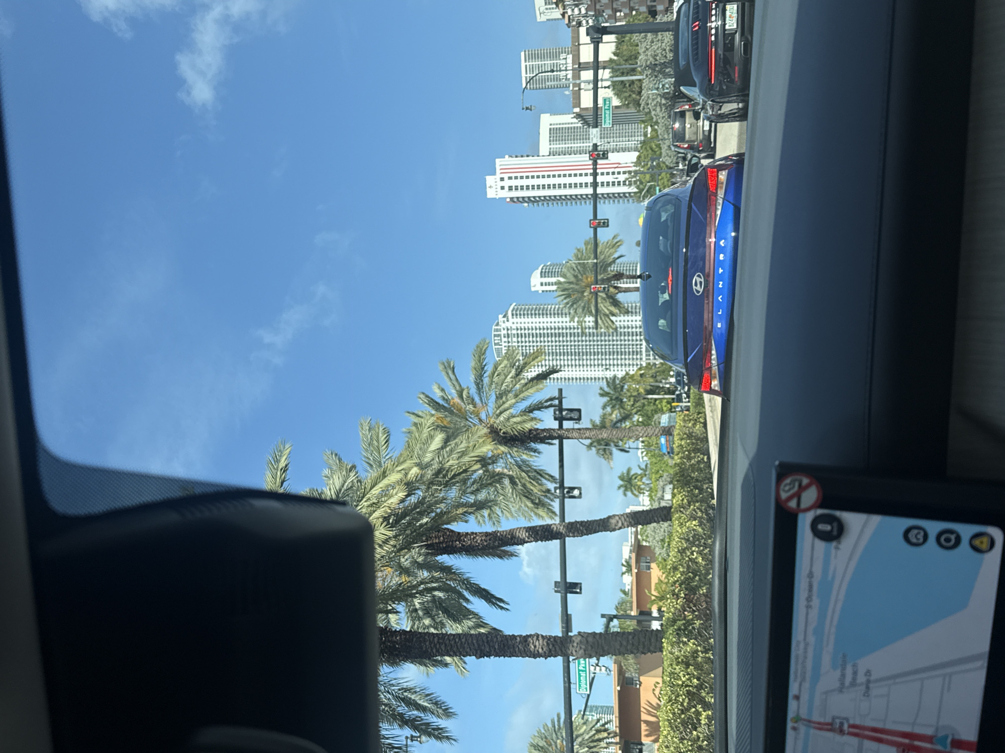 The image depicts a view from inside a car, seemingly in motion. The foreground features parts of the car's interior, including the dashboard and rearview mirror. A navigation screen is visible, displaying a map showing the car's location in Hallandale Beach, United States of America. The road network and streets named "Ocean Dr," "Hallandale Beach," and "Dune Dr" are discernible on the map. The car is positioned in a line of traffic, with a blue car directly ahead. Further beyond, modern high-rise buildings, presumably residential or commercial towers, are visible against a clear blue sky dotted with sparse clouds. Tall palm trees line the street, adding a tropical element to the urban landscape. The scene suggests a sunny day in a coastal city.