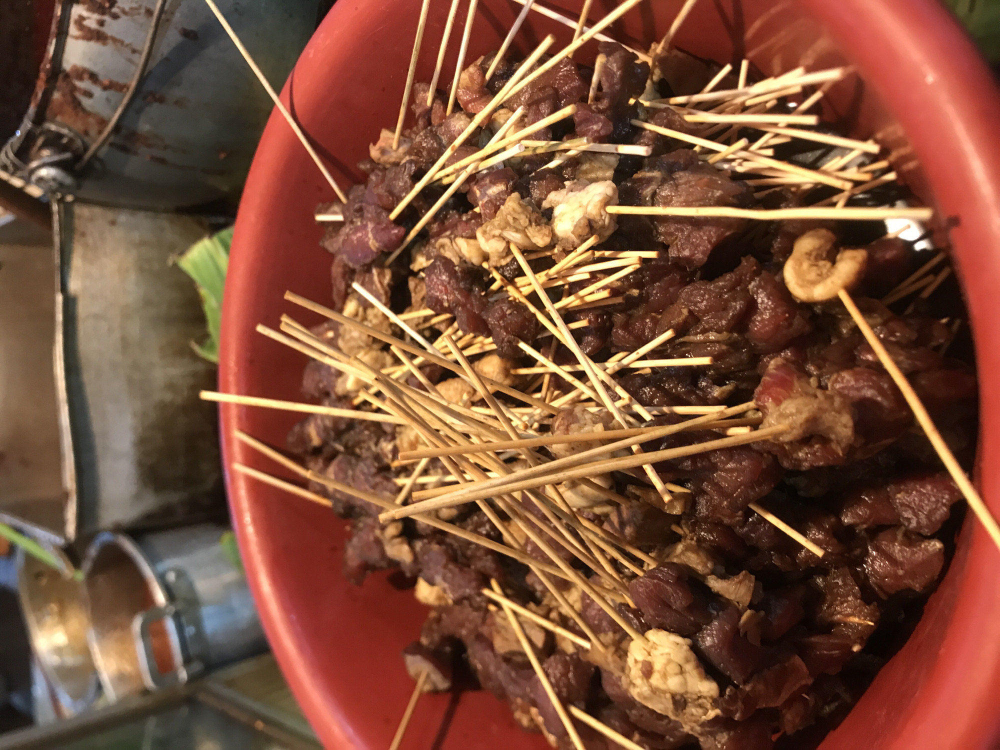 A close-up, high-angle shot captures a large, deep red plastic bowl overflowing with numerous uncooked meat skewers, commonly known as sate (satay), typical of Indonesian cuisine. The skewers are made of thin wooden sticks, each holding several irregular chunks of marinated meat. Most of the meat pieces are dark reddish-brown, while some lighter, more yellowish-white pieces, possibly fat or offal, are also visible. The skewers are piled densely within the bowl, some sticking out at various angles.
In the blurred background to the left, rustic and somewhat weathered metallic objects are partially visible, suggesting a functional or industrial setting, possibly a street food stall or a market kitchen. Elements like rusty metal surfaces and cylindrical containers contribute to this unrefined, utilitarian atmosphere. A hint of a green leaf can be seen behind these objects, adding a touch of natural color.
There are no people or visible text in the image. The lighting appears bright and even, consistent with daylight or strong artificial illumination, but the exact time of day or weather conditions cannot be determined from the indoor or covered setting. Given the location context of Cotpuuk, Indonesia, this image depicts a common local food preparation, likely ready to be grilled and served.