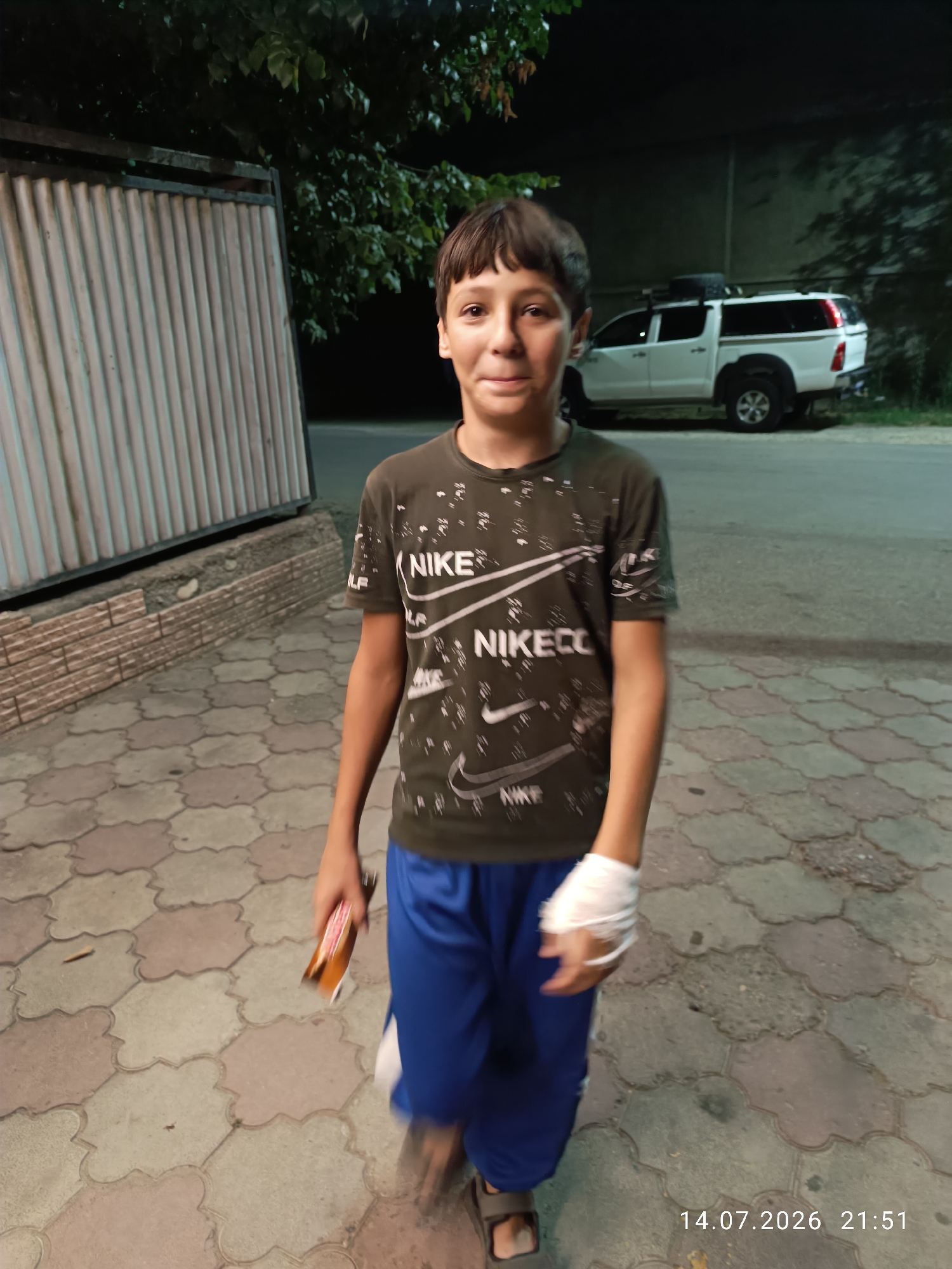 A young boy with short brown hair and a light complexion stands on a paved walkway at night. He is wearing a dark green t-shirt with white Nike branding and blue athletic shorts. His left hand is wrapped in a white bandage. He is holding a small, rectangular object, possibly a candy bar, in his right hand. Behind him, a white pickup truck with a camper shell and a spare tire on the roof is parked. A corrugated metal fence and trees are visible in the background. The timestamp on the image indicates the photo was taken on July 14, 2026, at 21:51. The location is identified as Qaskeleñ, Kazakhstan.