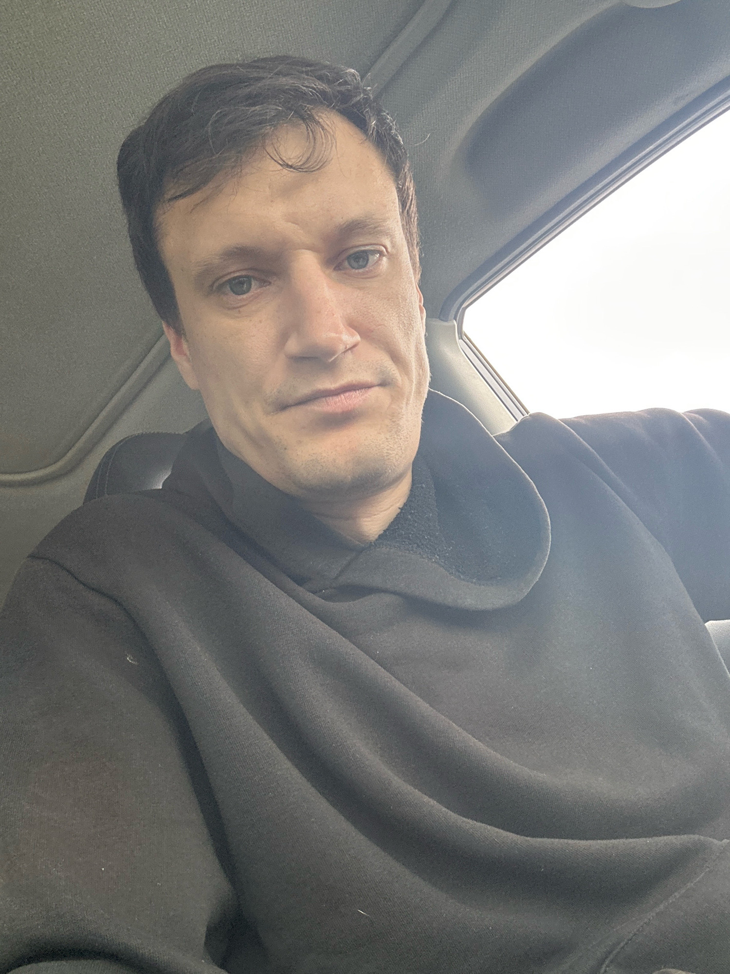 A close-up, low-angle, selfie-style shot captures a man in the driver's seat of a car, looking towards the camera. The man has short, dark brown hair and is wearing a black hooded sweatshirt. The interior of the car is visible in the background, with the headliner and a portion of the sunroof. The lighting suggests an overcast day. The image does not provide any visual information to indicate the location in the city of Makiv, Ukraine.