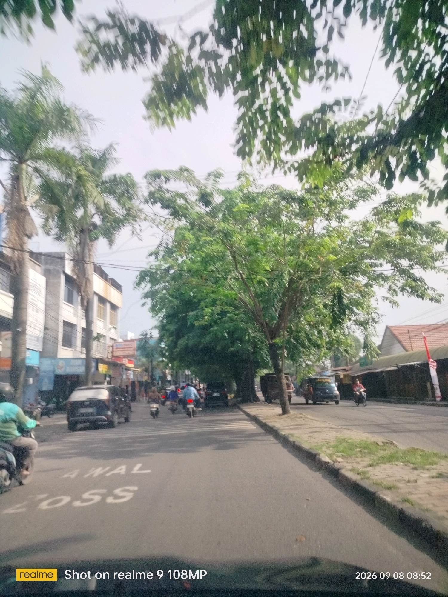 The image captures a street scene in the city of Medan, Indonesia. The foreground shows a road with white markings, partially obscured by what appears to be the dashboard of a vehicle. Two prominent white markings on the road are visible: "AWAL" and "LOSS". Several vehicles, including motorcycles and cars, are driving on the road. On the left side of the street, there are multi-story buildings and palm trees lining the sidewalk. On the right, a row of shops with awnings is visible. The sky is overcast, and the overall atmosphere suggests an ordinary day in an urban environment. The timestamp on the bottom right corner indicates the photo was taken on September 8, 2026, at 08:52.