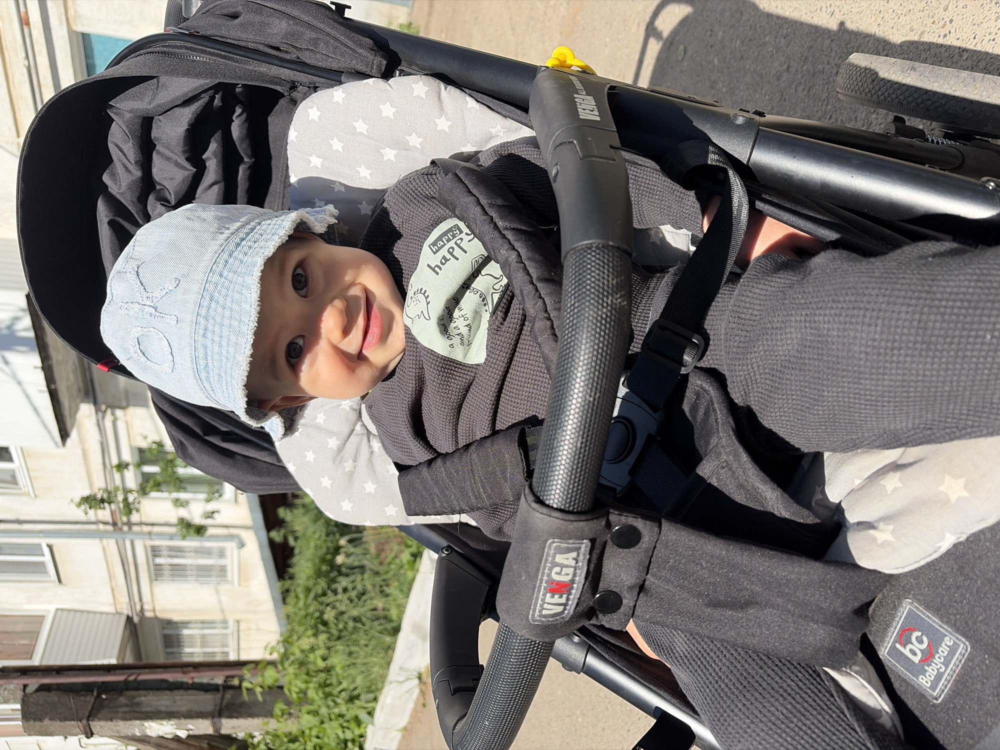 A baby is positioned in a black stroller with a gray and white star-patterned liner. The baby is wearing a light blue denim hat and a dark gray waffle-textured outfit with a "happy happy" patch on the chest. The stroller has a black handle with the "VE GA" brand name visible. Another brand, "bc Babycare", is also visible on the stroller. The background shows a light-colored building with several windows and some greenery. The location appears to be in an urban area of Chaikovsky, Russia.