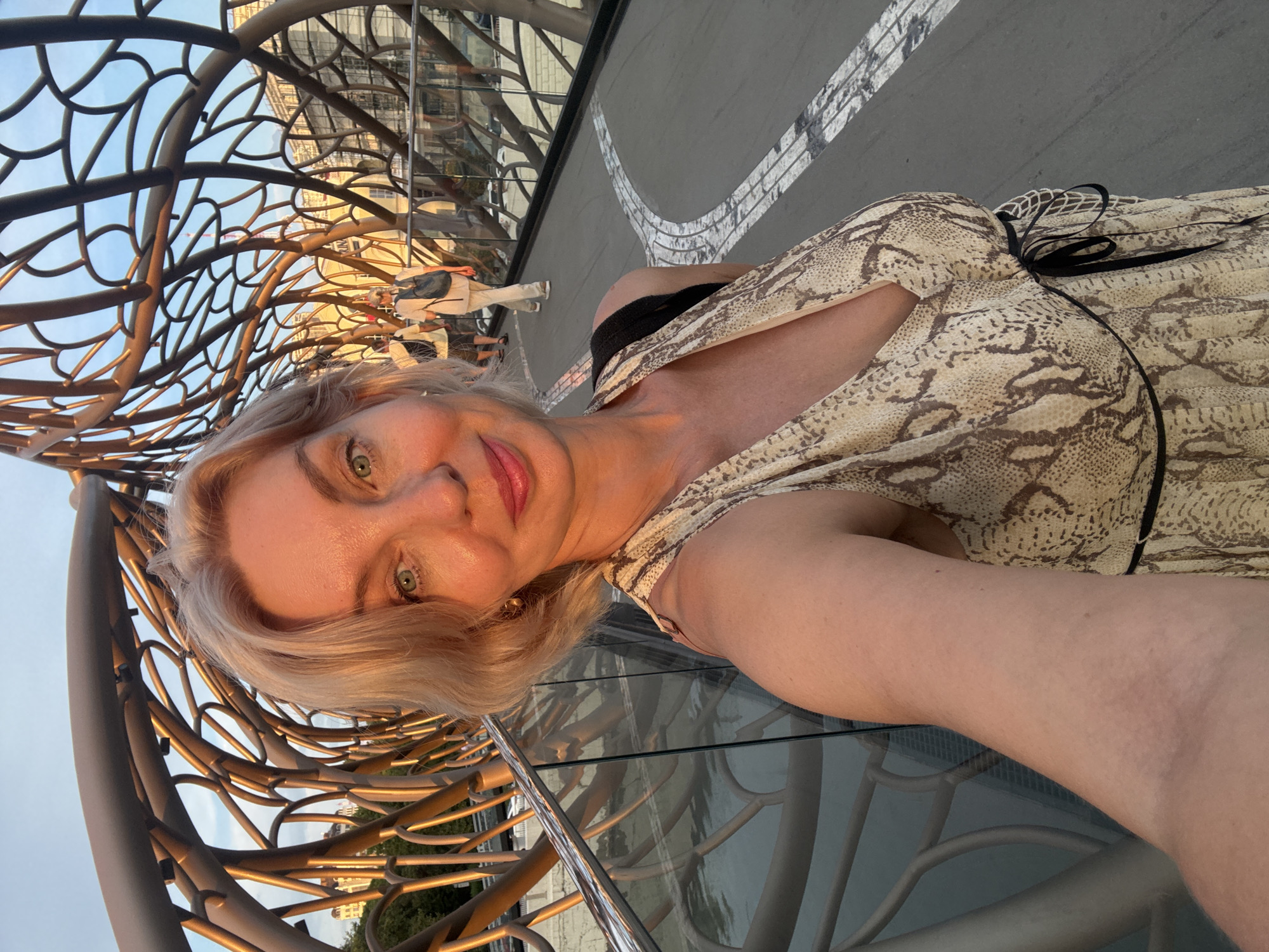A blonde woman with green eyes and a snakeskin-print dress takes a selfie on a bridge with a unique, organic-looking metal structure. The bridge appears to be in a city, with buildings visible in the background. The sky is clear and blue, and the lighting suggests it is either sunrise or sunset. The woman is smiling and looking at the camera. In the background, other people can be seen walking on the bridge. The bridge has glass railings and a light-colored walkway with decorative white lines. This is likely the Bridge of Love in Sochi, Russia, known for its distinctive architectural design.