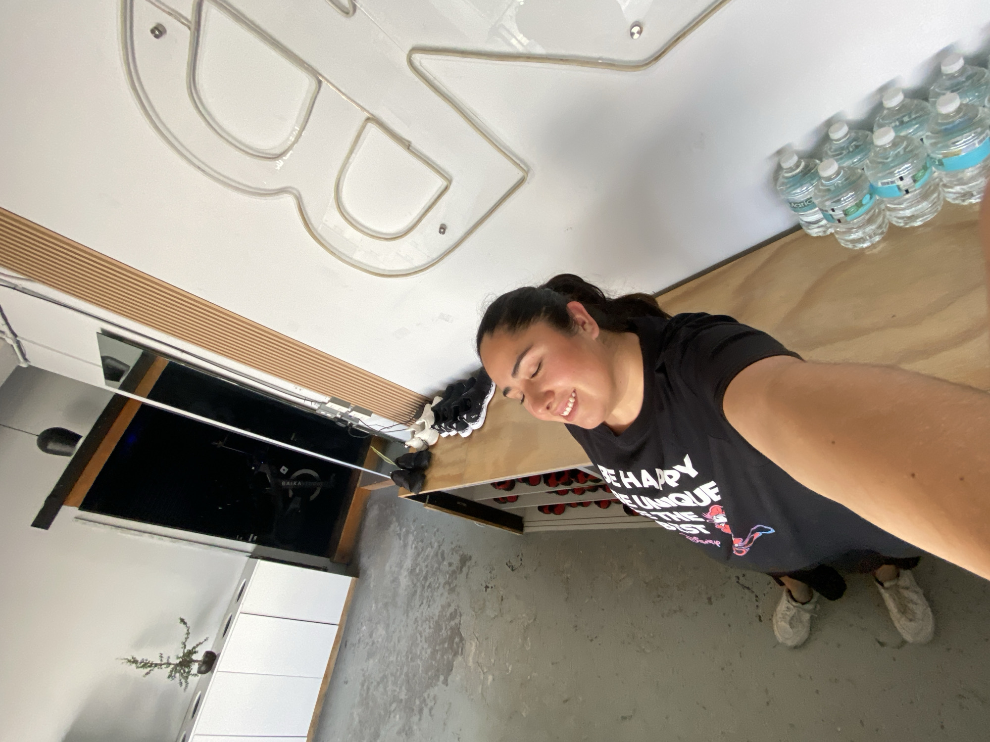 The image, likely taken in Puebla, Mexico, captures a young woman in a moment of apparent happiness, smiling with her eyes closed. She has dark hair pulled back in a ponytail and is wearing a black t-shirt that reads "BE HAPPY BE UNIQUE BE THE BEST" with a faint "Disney" logo below. She is wearing light-colored sneakers on a grey, somewhat worn concrete floor.
The setting appears to be a modern indoor space, possibly a gym or a fitness studio. On a high white wall or ceiling, a large, illuminated or outlined logo, resembling stylized letters such as "BR", is visible. Below this, a row of plastic water bottles with blue labels, some partially showing the brand "Maria", are neatly arranged on a ledge. Beneath the woman's head, a wooden shelf holds a pair of black and white sneakers. Below this shelf, a storage unit filled with numerous red athletic shoes, possibly cycling shoes, are organized in cubbies. To the left, a large reflective surface, which could be a mirror or dark glass, shows a partial reflection of another room, including a small potted plant on a white cabinet and the faint text "BAIKAL HOUSE". The overall impression is that of a functional, contemporary environment related to health or fitness.