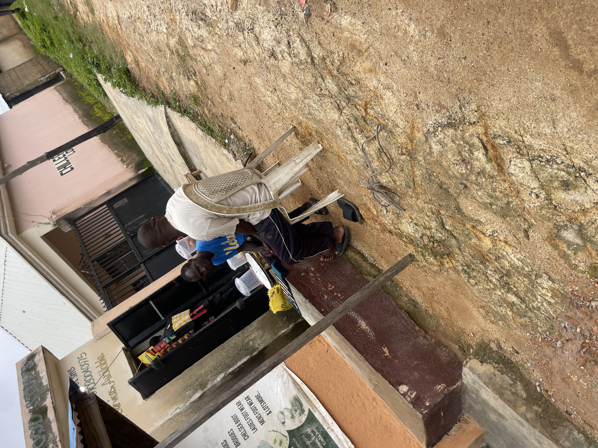 Two men are seated on plastic chairs in front of a small shop on a dusty, uneven path in Jos, Nigeria. The man on the left wears a blue t-shirt with white lettering and black track pants. He is looking towards the viewer. The man on the right wears a light-colored t-shirt and dark track pants, and he is looking away from the viewer. A white plastic table with some containers and a yellow bag is situated between them. In the background, a building with a light-colored facade and a dark-colored entrance with metal bars is visible, along with a pink wall bearing the word "CHILLE" in black lettering. The ground is a mix of dirt, sand, and small stones. A pile of tangled wires lies on the ground to the right of the chairs. A sign is partially visible on the left side of the image, listing various footwear items and contact information. The overall scene depicts a casual outdoor setting on a potentially overcast day.
