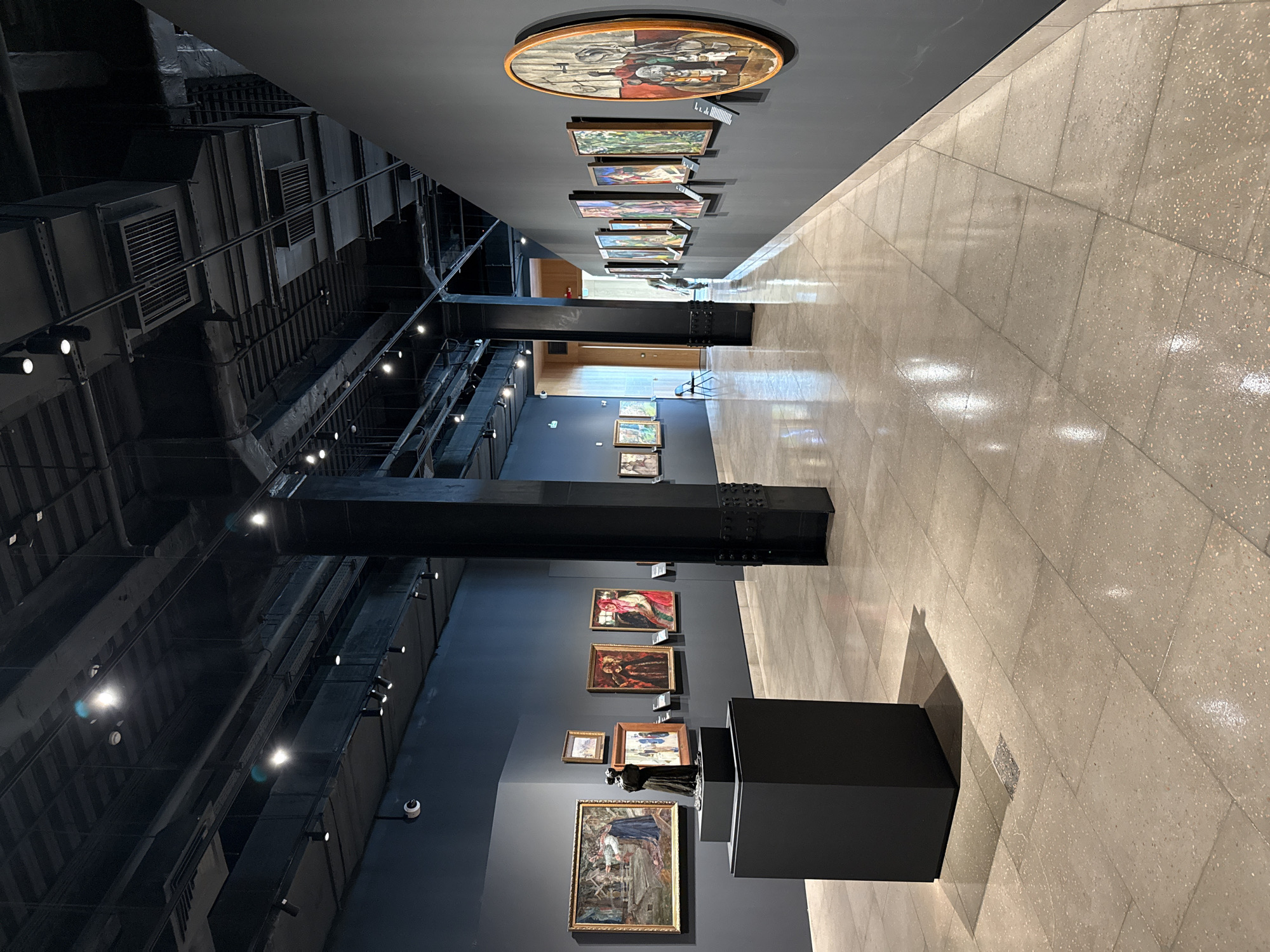 The image shows the interior of an art museum gallery. The walls are painted dark gray, and the ceiling is black with exposed ductwork and track lighting. The floor is made of large, light-colored tiles with a speckled pattern.
On the left wall, a series of paintings are displayed. In the foreground, a dark gray pedestal supports a dark sculpture of two figures. The paintings on the left wall appear to be figurative or narrative.
The gallery extends into the distance, with more paintings displayed on the right wall. These paintings are varied in size and framing, and some are circular. The lighting is focused on the artwork, creating spotlights and shadows. The overall impression is one of a modern, well-lit exhibition space. There are no people clearly visible in the immediate foreground or midground of the image, though a figure can be seen in the far distance towards the right.
This image does not provide any information about the city of Perm, Russia, or any specific location.