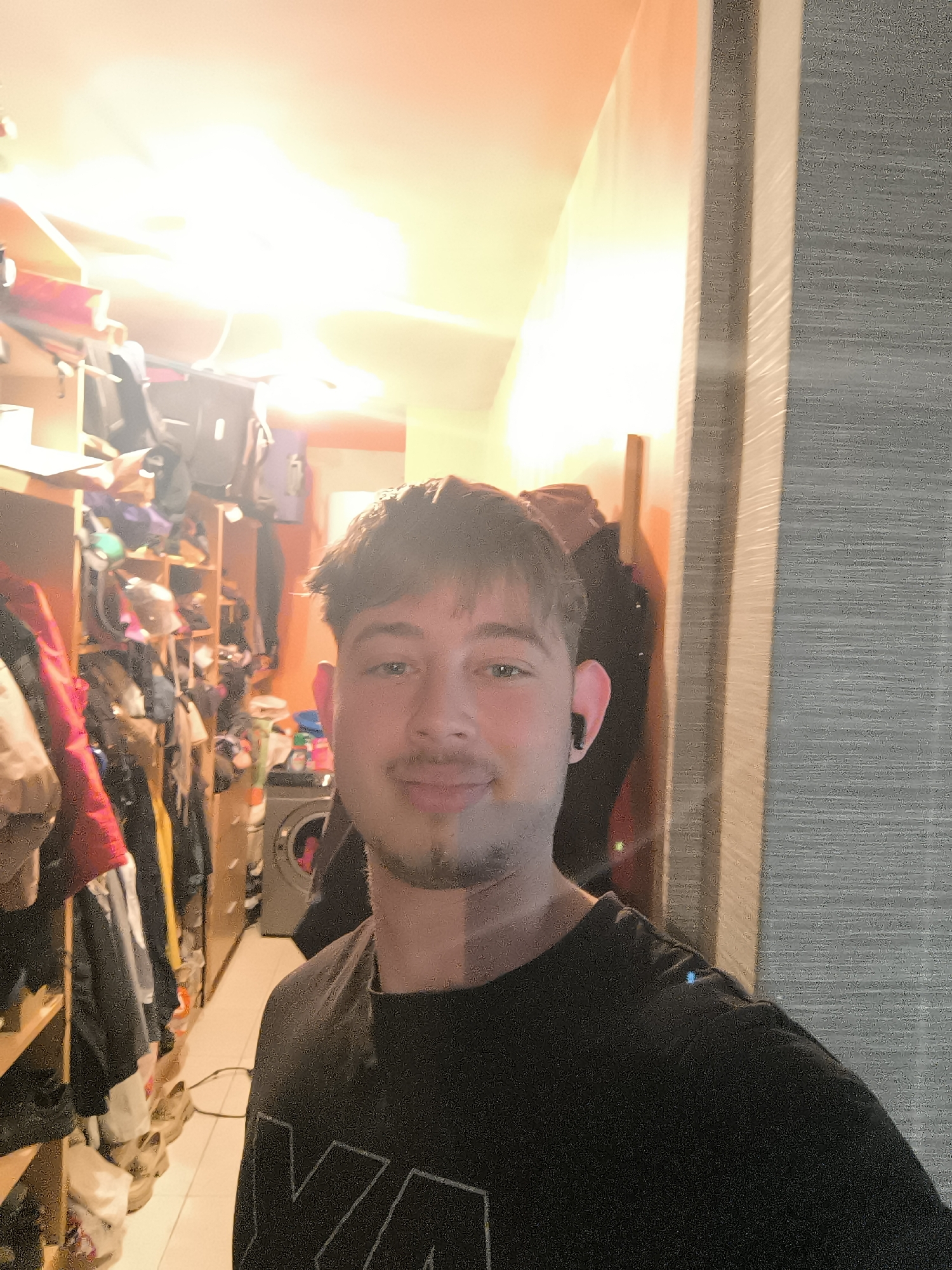 A medium shot captures a young man with fair skin, brown hair, and a short beard, wearing a black t-shirt and a black wireless earbud in his left ear. He is smiling softly at the camera. Behind him, to the left, is a brightly lit walk-in closet cluttered with clothing, shoes, and bags. A washing machine is visible at the end of the closet aisle. To the right of the man, there is a gray textured wall with a light-colored door frame. The image appears to be taken indoors. There is no information in the image that would indicate that this is in Sofia, Bulgaria.