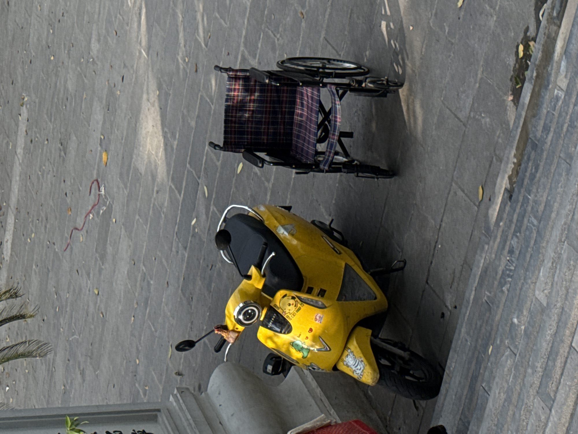 In Hà Nội, Vietnam, a yellow scooter with cartoon stickers and a folded wheelchair rest on a gray stone pavement adjacent to a stone staircase. Palm fronds are visible on the lower left.