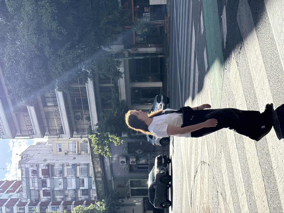 An individual is depicted mid-stride, walking left to right across a pedestrian crosswalk on an urban street in Buenos Aires, Argentina. The person is a female with shoulder-length brown hair, wearing a white t-shirt, dark wide-leg pants, and dark shoes, with a black fanny pack or sling bag worn across her chest. Strong sunlight illuminates her from the right and behind, creating a halo effect on her hair and casting distinct shadows on the asphalt.
The street features white zebra-stripe markings for the crosswalk and a segment of a green-painted lane to the right. In the background, parked or slowly moving vehicles are visible, including a dark grey SUV on the left and a silver SUV further right, both facing left.
The surrounding environment consists of multi-story buildings characteristic of Buenos Aires architecture. On the left, an older, lower structure with a distinct purplish and green facade and multiple air conditioning units is topped by a taller residential building with numerous balconies. On the right, a modern-style white multi-story building features large windows and balconies with horizontal railings. Its ground floor appears to house commercial establishments, with a partial "express" sign visible. Green foliage from trees lines parts of the street. The sky above is clear with some scattered clouds, indicating a bright, sunny day. A prominent lens flare from the sun traverses the upper right portion of the scene.