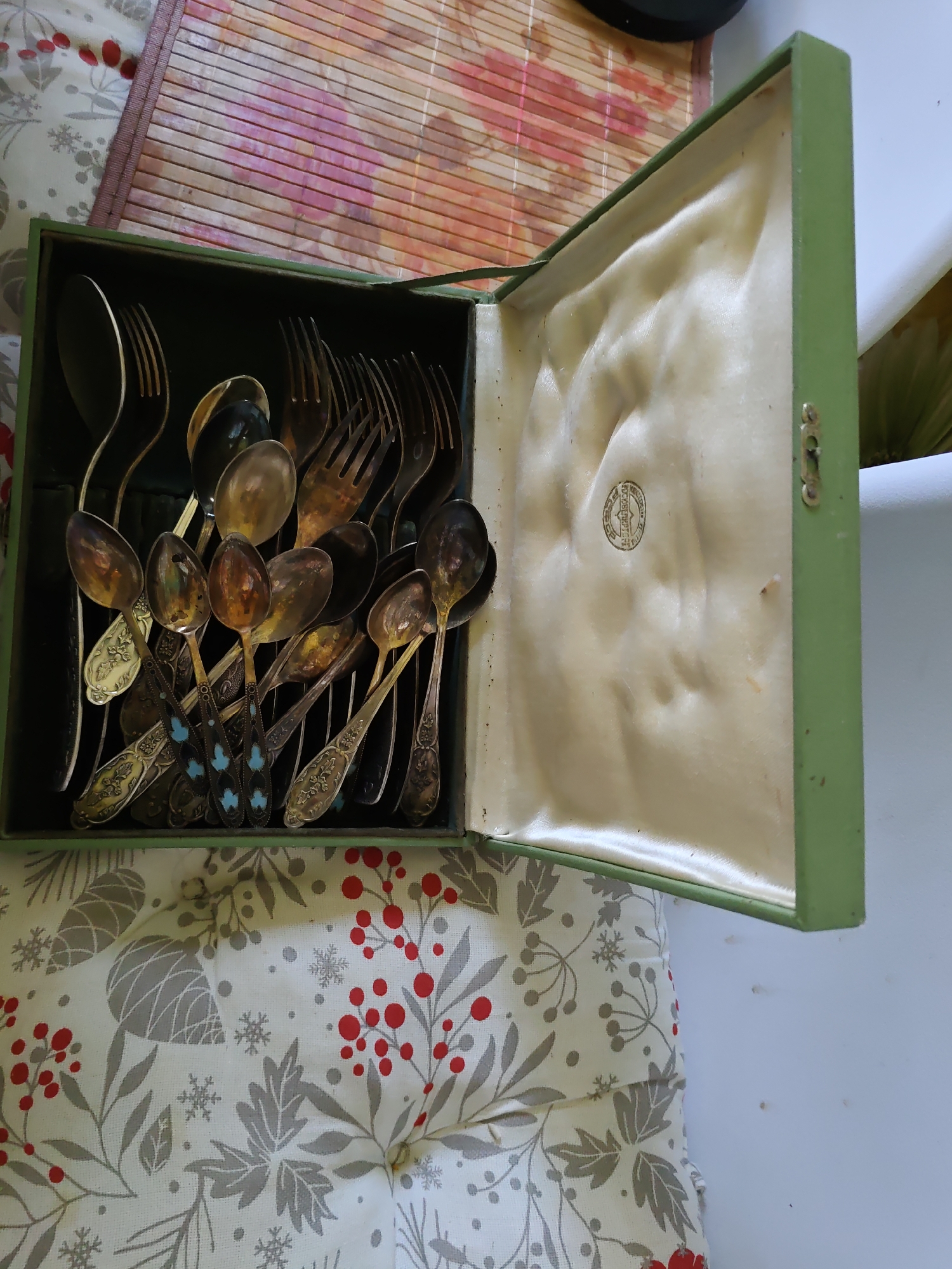 The image shows an open green box containing an assortment of antique silverware, including spoons and forks. The silverware is tarnished and ornate, with some pieces featuring blue and white patterns on their handles. The box is lined with a cream-colored satin fabric, which has a subtle quilted pattern and a circular emblem pressed into it. The box is placed on a surface covered with a festive fabric featuring red berries, grey leaves, and snowflakes. In the background, a portion of a bamboo mat is visible. The image is taken from a slightly elevated angle, looking down into the box. There are no people or animals present in the image. The lighting suggests an indoor environment. The context of Rostov-on-Don, Russia is not visually evident in the image itself.
