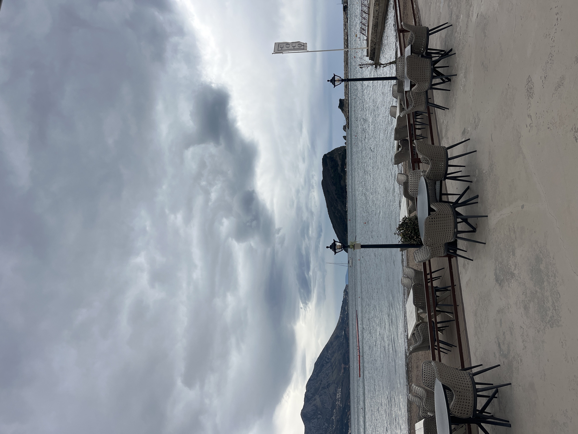 The image captures a seaside scene in Budva, Montenegro. A row of empty cafe tables and chairs lines a concrete terrace overlooking the Adriatic Sea. The sky is overcast with heavy, grey clouds. In the distance, mountains rise from the sea. A flag with glasses printed on it is placed near a lamp post next to the water.