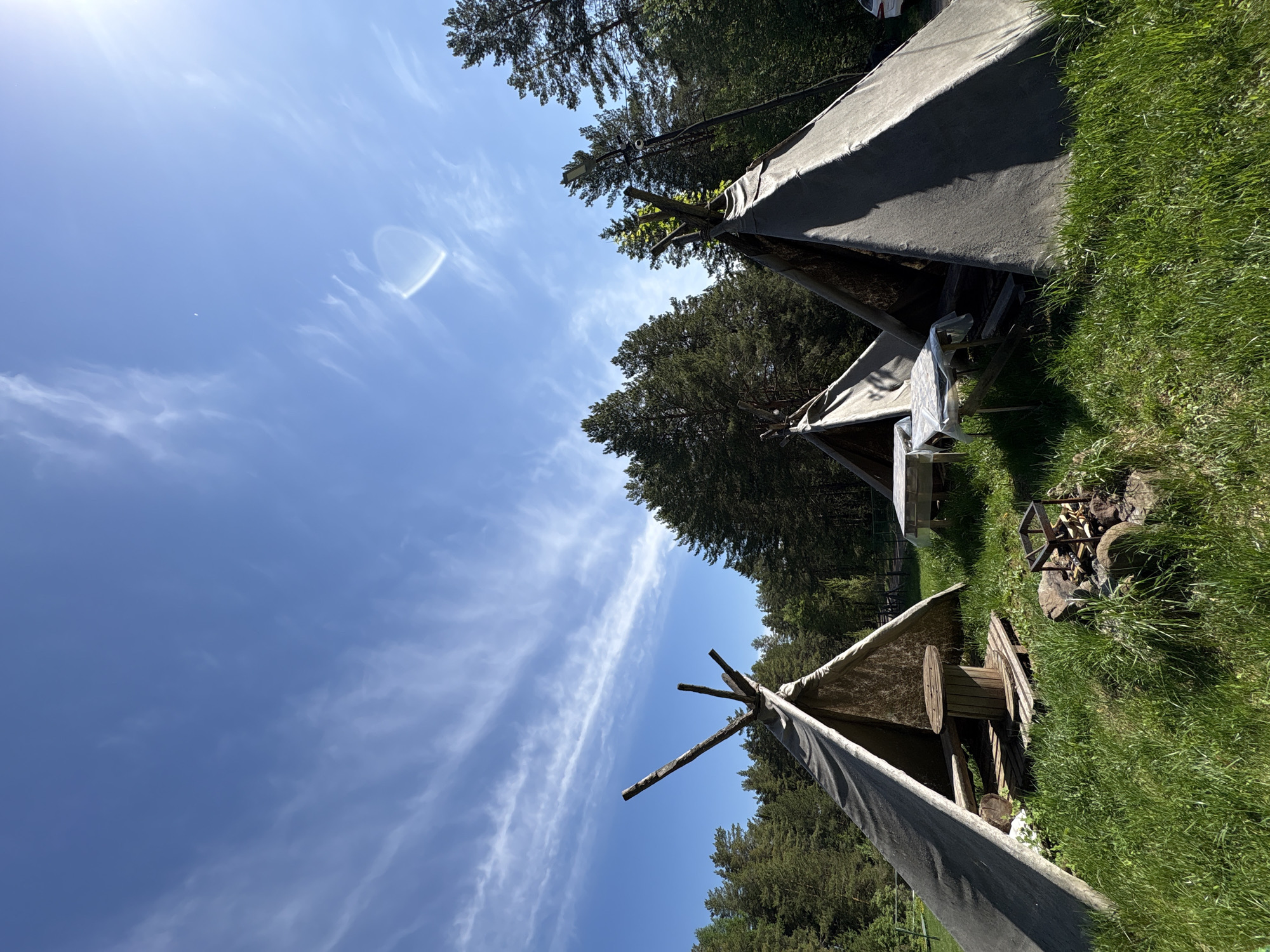 The image shows a campsite in Прикамский, Russia. Three teepees stand on a grassy area, surrounded by trees. The sky is blue with some wispy clouds. A fire pit is visible near one of the teepees, with wood stacked inside. One of the teepees has a large wooden spool inside.