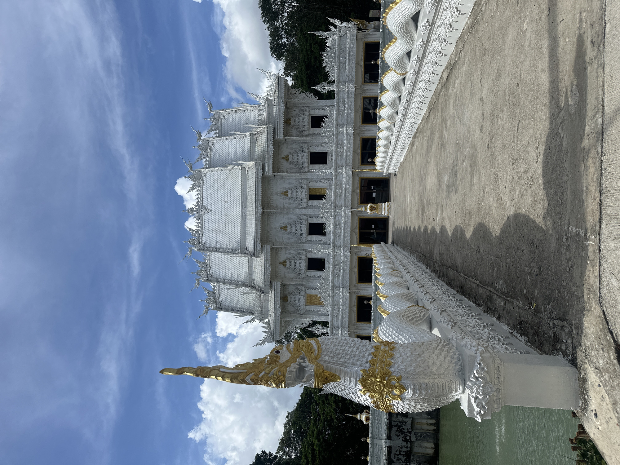 This is a frontal, eye-level, medium-range, full-shot photograph of a white, ornate temple with intricate architectural details, set against a bright blue sky with scattered white clouds. The temple is the main subject, dominating the upper portion of the frame. A wide, concrete walkway leads directly to the temple's entrance. Flanking the walkway is a decorative white railing topped with a repeating pattern of white, scaled serpents with gold accents on their heads and collars. To the left of the walkway, a body of green water is visible, with a large, white, serpentine sculpture, also with gold accents, positioned prominently in the foreground. The temple itself features multiple tiered roofs with pointed, ornate finials. The facade is adorned with elaborate carvings and decorative elements, with arched windows and doorways. Gold accents are visible on some window frames and decorative motifs. The overall impression is one of grandeur and spiritual significance. The scene is set outdoors, with lush green trees visible in the background behind the temple and to the sides, suggesting a natural or park-like setting. The lighting indicates it is daytime, likely late morning or early afternoon, with strong shadows cast on the walkway. The location is identified as Thep Krasatti, Thailand. There are no people or animals visible in the image.